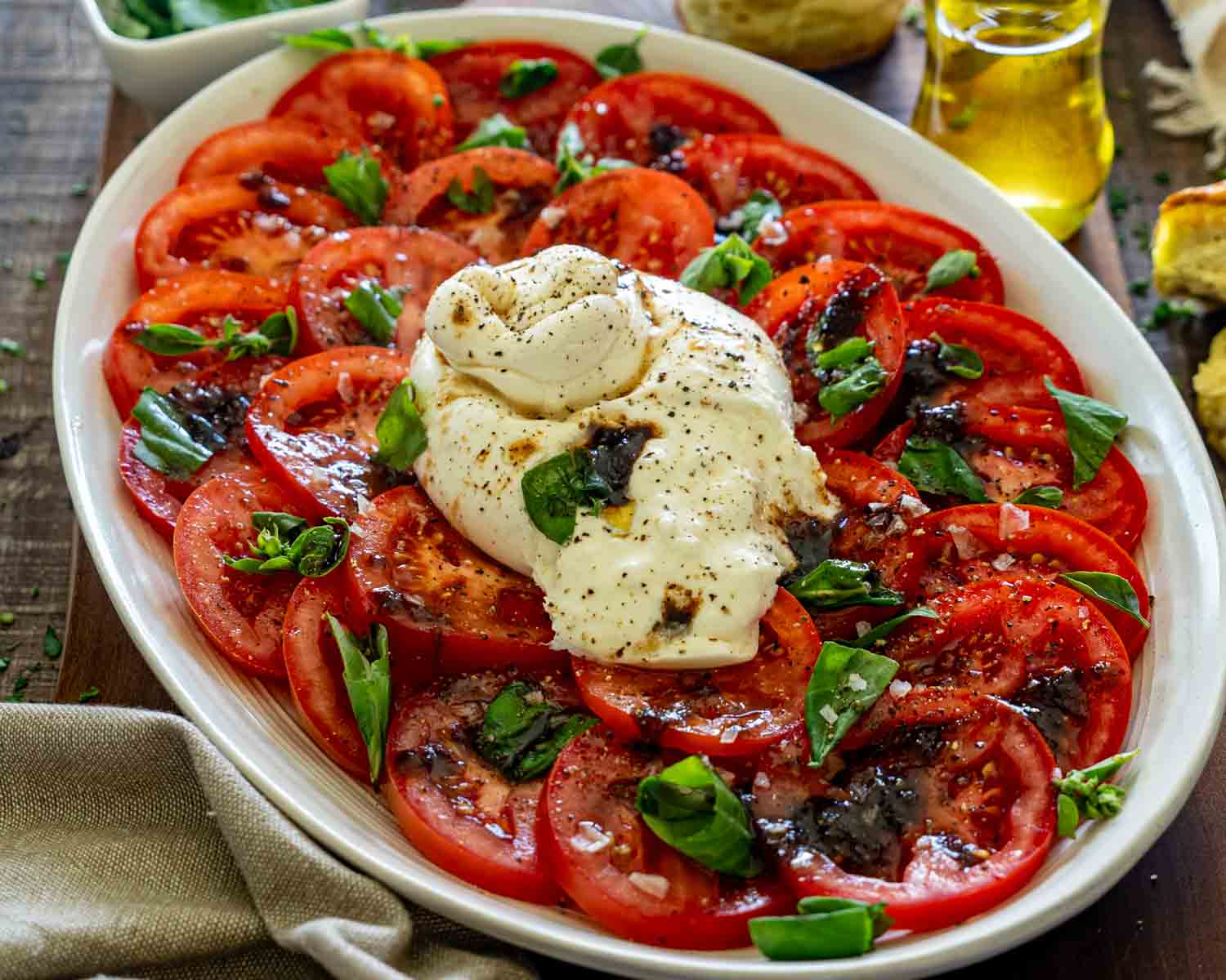 Fresh tomato burrata salad arranged on oval platter with basil leaves, balsamic glaze, flaky salt, and creamy burrata cheese.