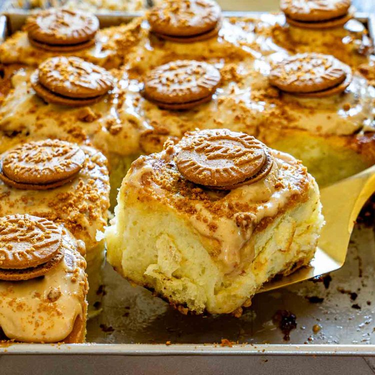 Close-up of a fluffy Biscoff cinnamon roll lifted from the pan, showcasing swirls, creamy frosting, and crunchy cookie topping.