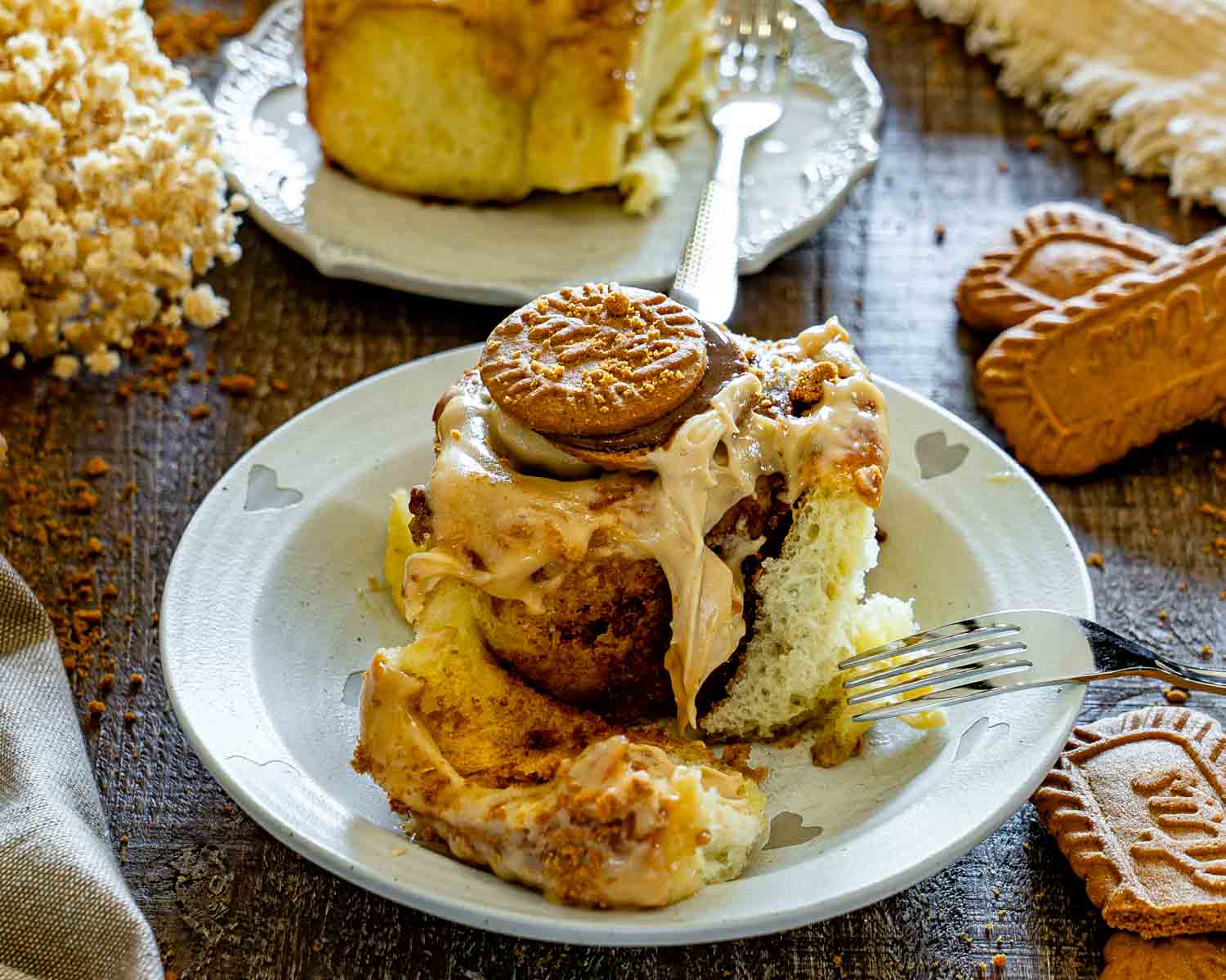 Soft Biscoff cinnamon roll with a bite missing, topped with creamy frosting and cookie, served on a plate with fork.