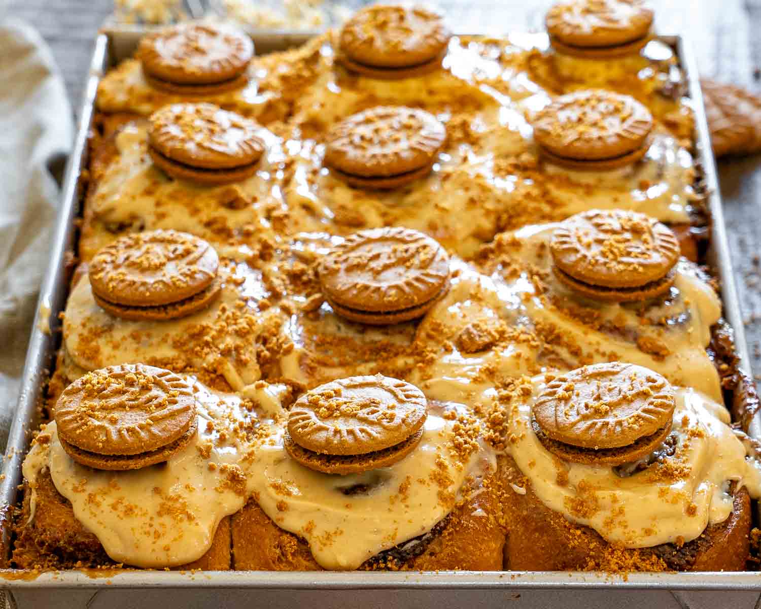 Freshly baked Biscoff cinnamon rolls covered with creamy Biscoff frosting, crushed cookies, and whole Biscoff biscuits in a metal baking pan.