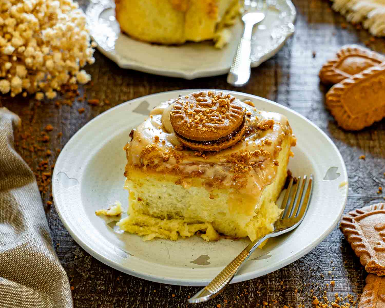Thick slice of Biscoff cinnamon roll served on a plate with creamy frosting, cookie topping, and another roll blurred behind. (