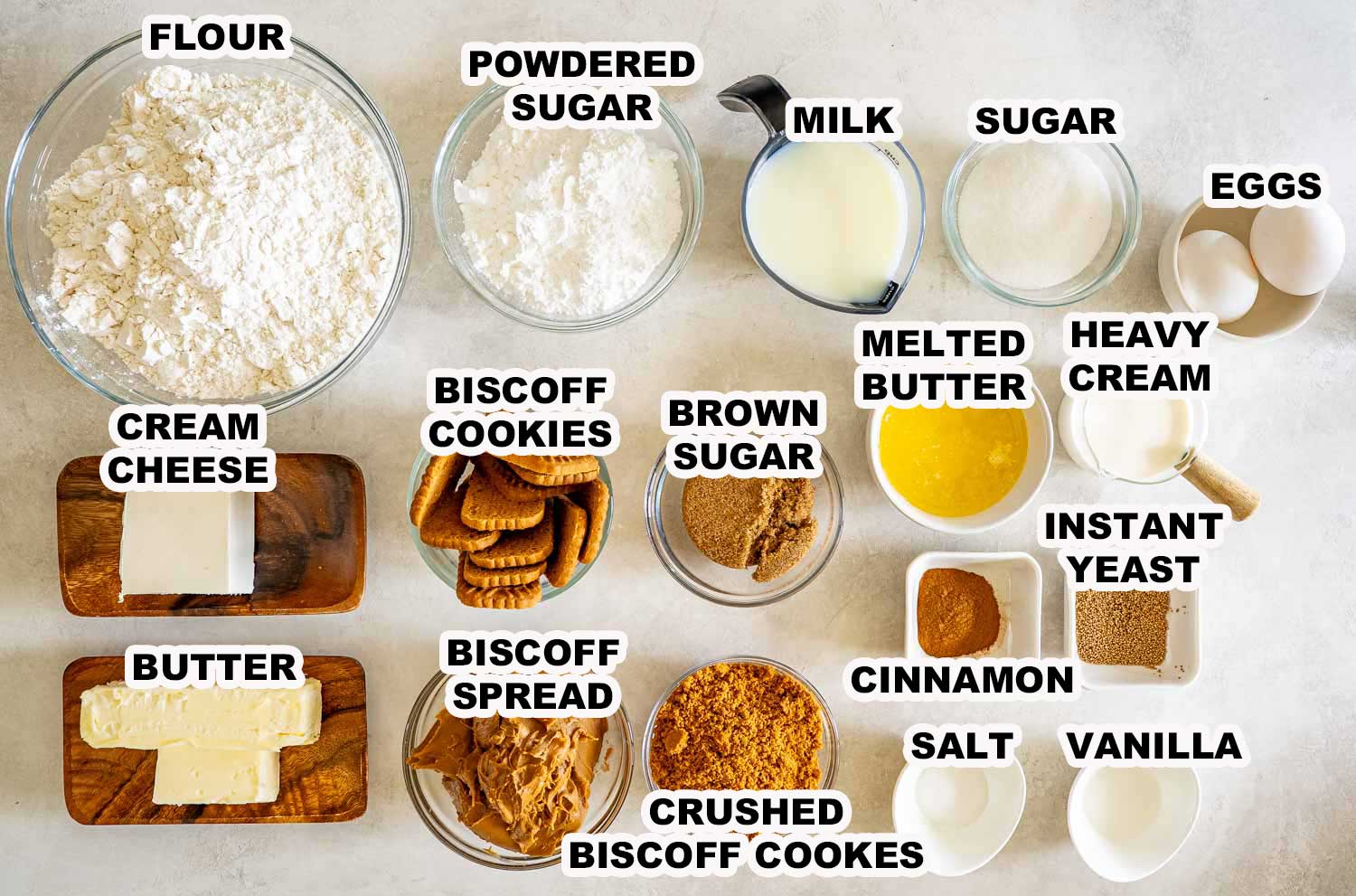 Overhead view of ingredients for Biscoff cinnamon rolls including flour, Biscoff spread, cookies, butter, cream cheese, yeast, cinnamon, and sugar.