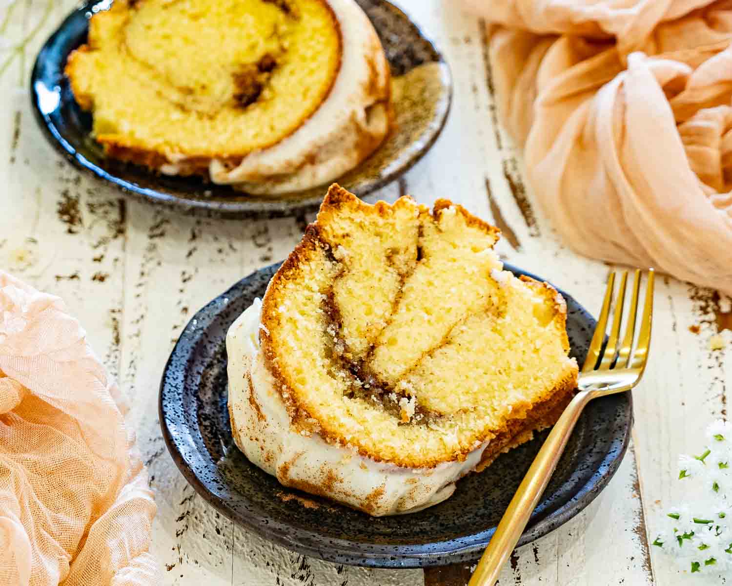 Thick slice of cinnamon roll cream cheese pound cake topped with cream cheese glaze, served on a black plate with a gold dessert fork.