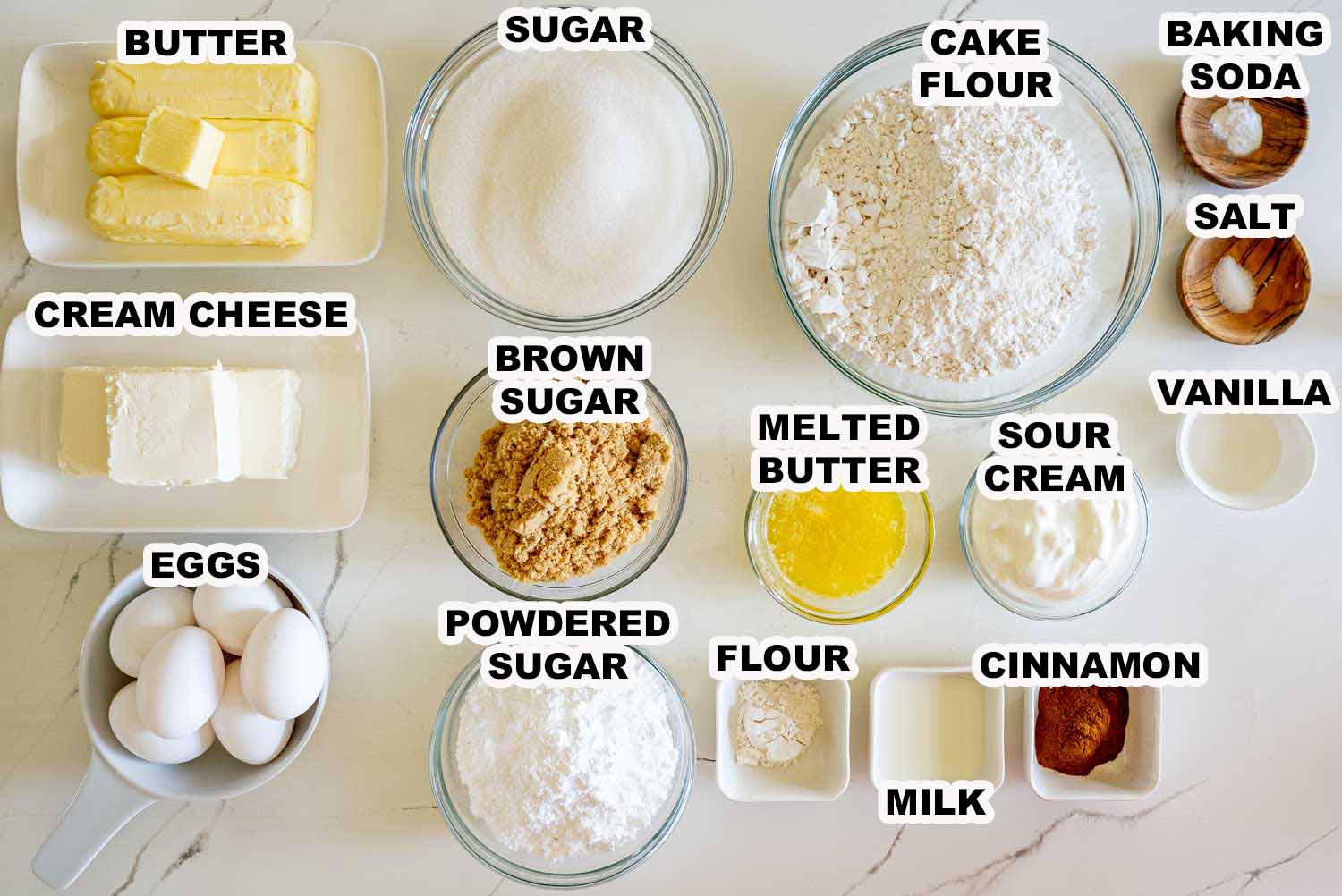 Overhead view of cinnamon roll cream cheese pound cake ingredients neatly arranged in bowls, labeled and ready for baking on a marble countertop.