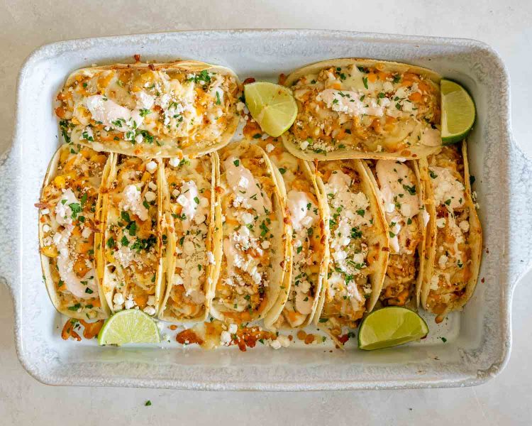 Creamy street corn chicken tacos arranged in a baking dish with melted cheese, creamy sauce, cilantro, feta, and lime wedges.