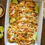 Overhead view of a baking dish packed with creamy street corn chicken tacos, crumbled feta, cilantro, sauce, and lime wedges.