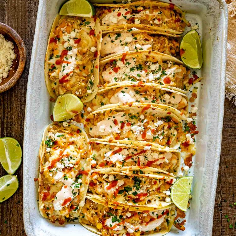 Overhead view of a baking dish packed with creamy street corn chicken tacos, crumbled feta, cilantro, sauce, and lime wedges.