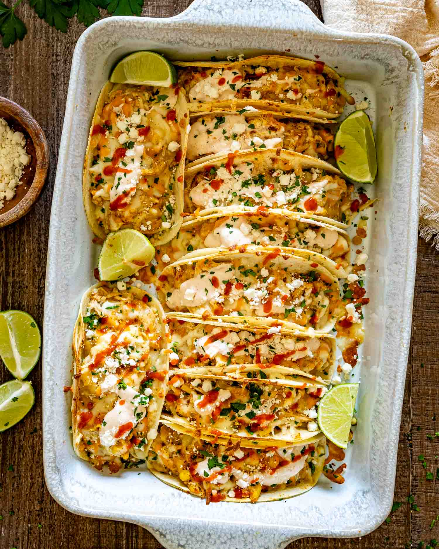 Overhead view of a baking dish packed with creamy street corn chicken tacos, crumbled feta, cilantro, sauce, and lime wedges.