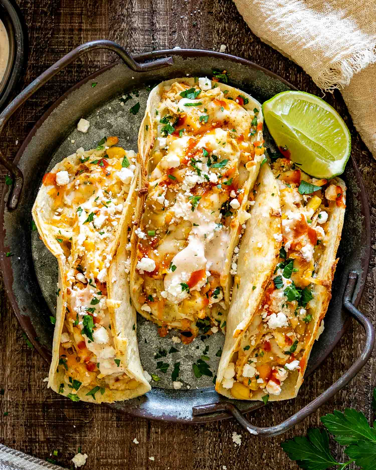 Overhead view of three creamy street corn chicken tacos topped with feta, cilantro, spicy sauce, and a fresh lime wedge.