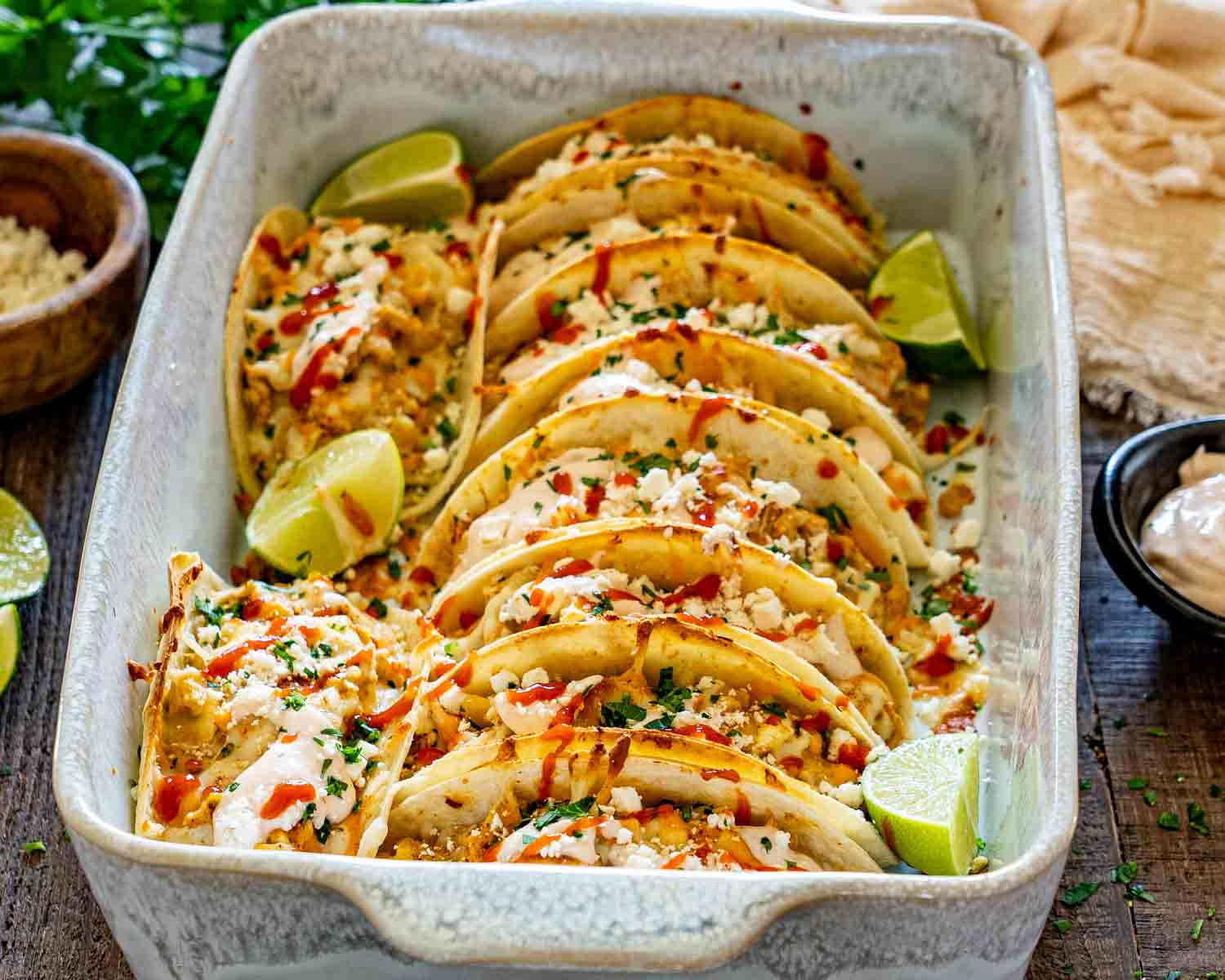 Close-up of creamy street corn chicken tacos arranged in a baking dish with feta, cilantro, hot sauce, and lime wedges.