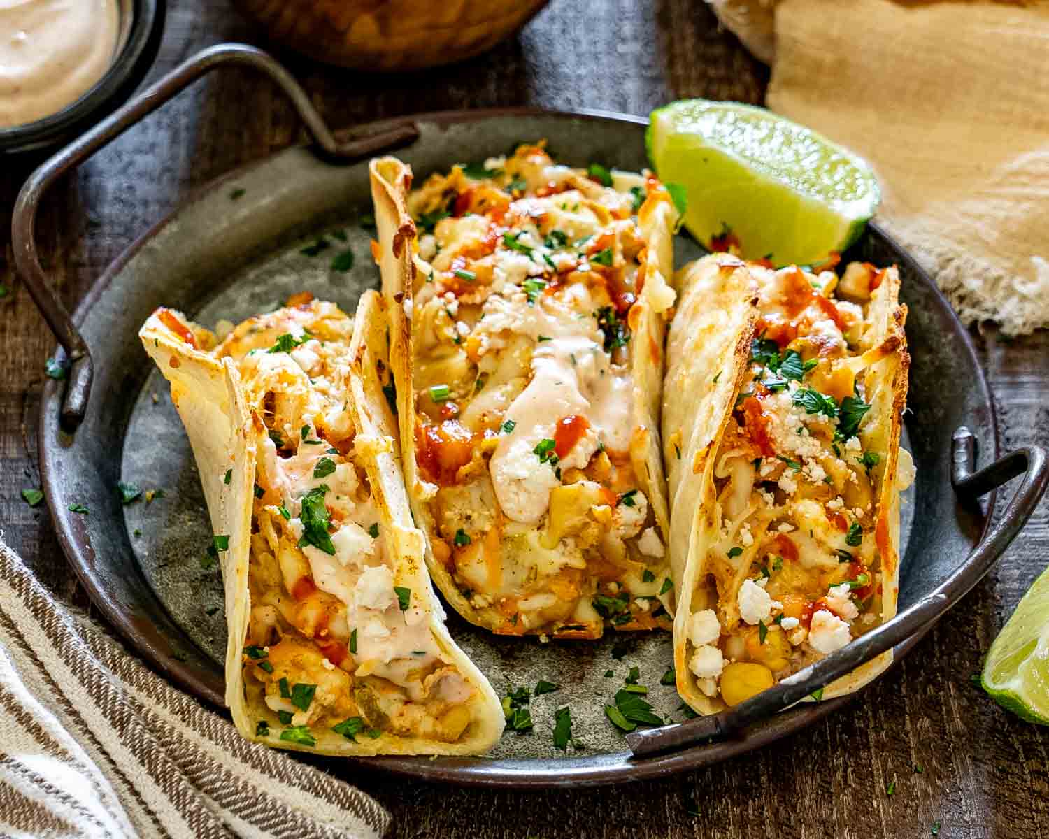 Three creamy street corn chicken tacos served on a rustic metal platter with crumbled feta, cilantro, spicy sauce, and lime.