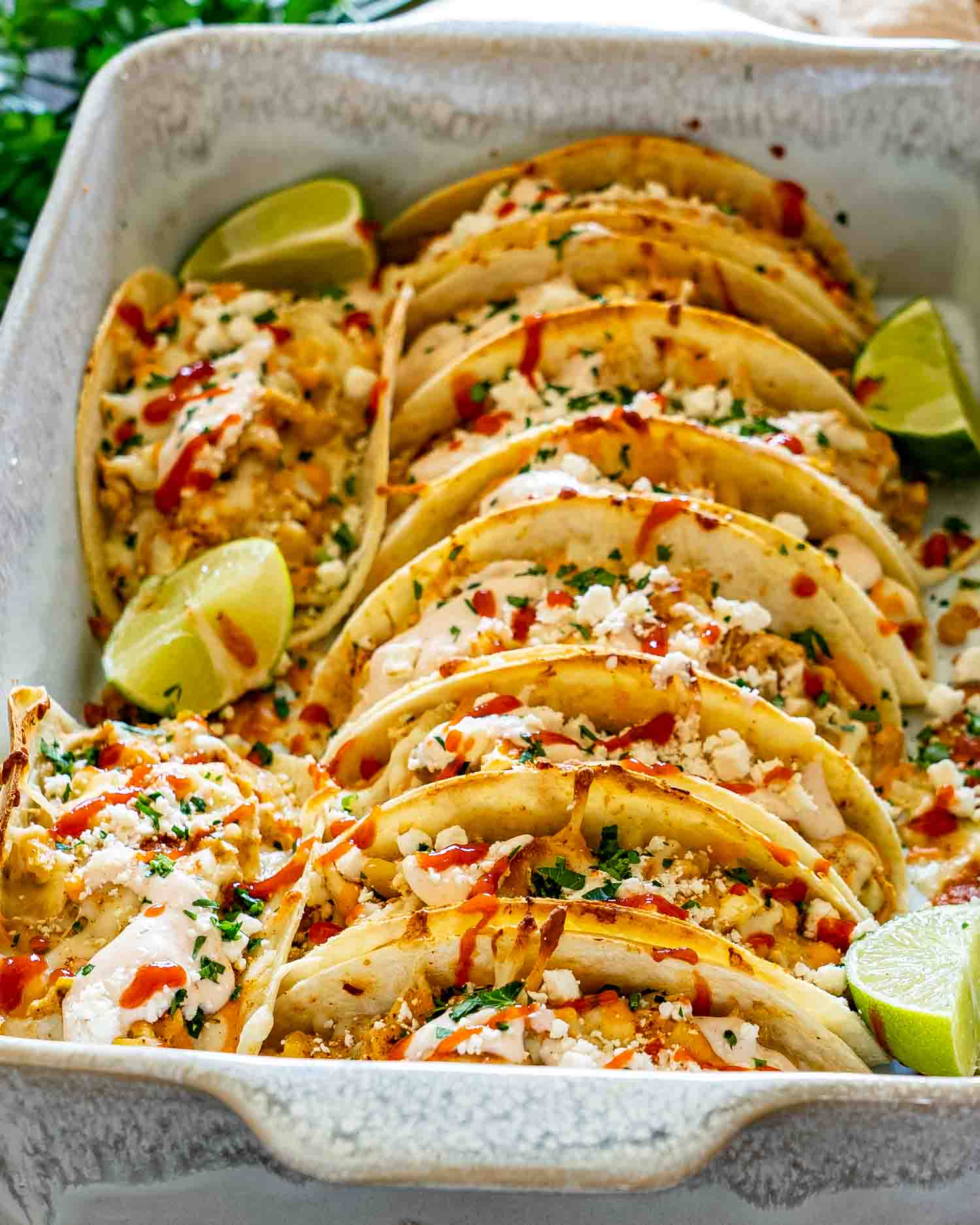 Close-up of creamy street corn chicken tacos arranged in a baking dish with feta, cilantro, hot sauce, and lime wedges.