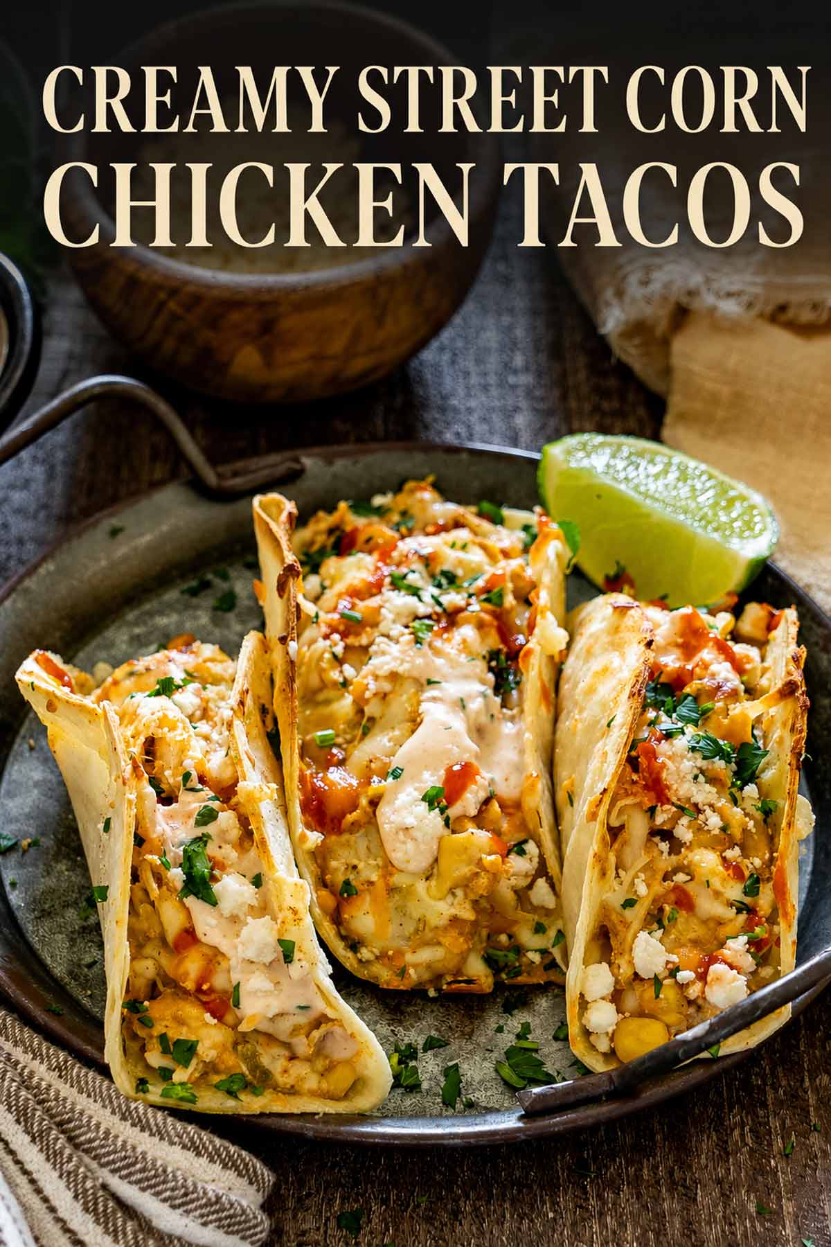 Pinterest image featuring three creamy street corn chicken tacos with melted cheese, spicy sauce, cilantro, feta, and lime.