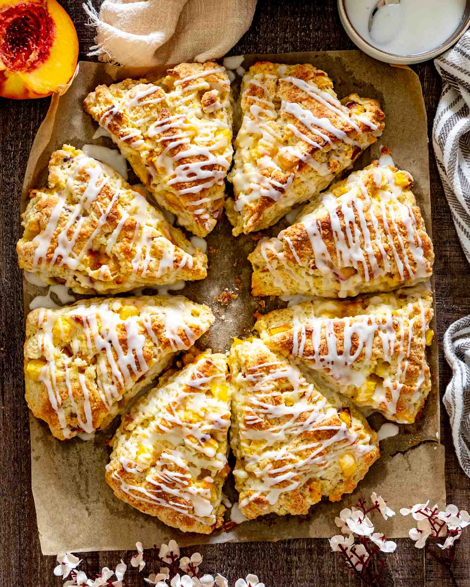 Overhead view of eight golden peach scones drizzled with vanilla glaze and arranged on parchment with fresh peach halves.