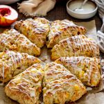 Golden peach scones drizzled with vanilla glaze and arranged on parchment beside fresh peaches and delicate white flowers.