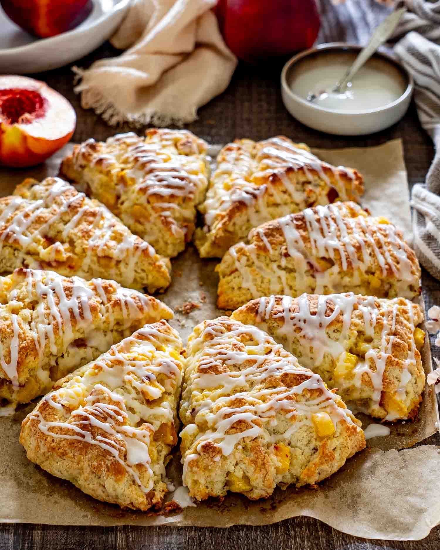 Golden peach scones drizzled with vanilla glaze and arranged on parchment beside fresh peaches and delicate white flowers.