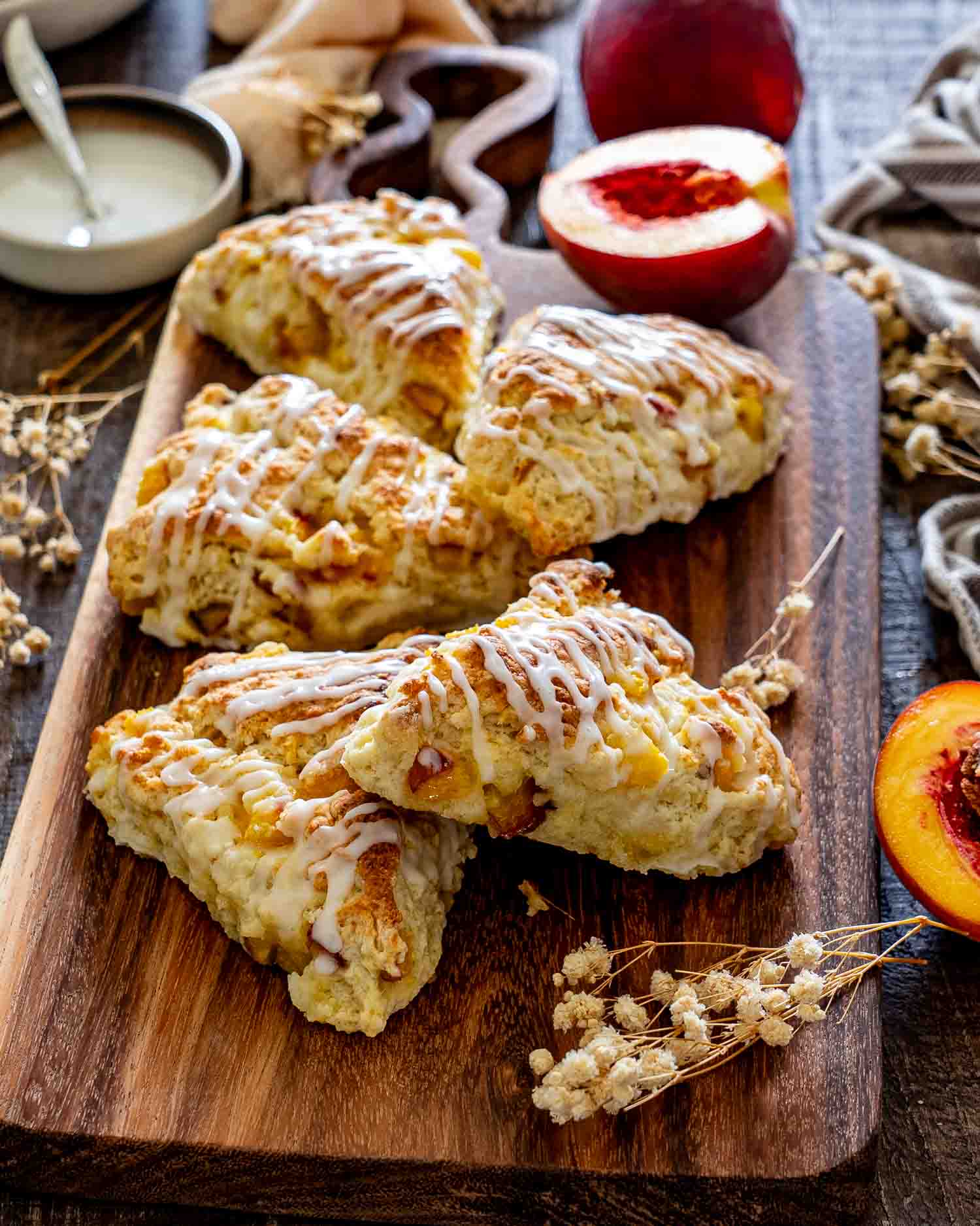 Glazed peach scones stacked on a rustic wooden board with fresh peaches, icing, linens, and delicate dried flowers.