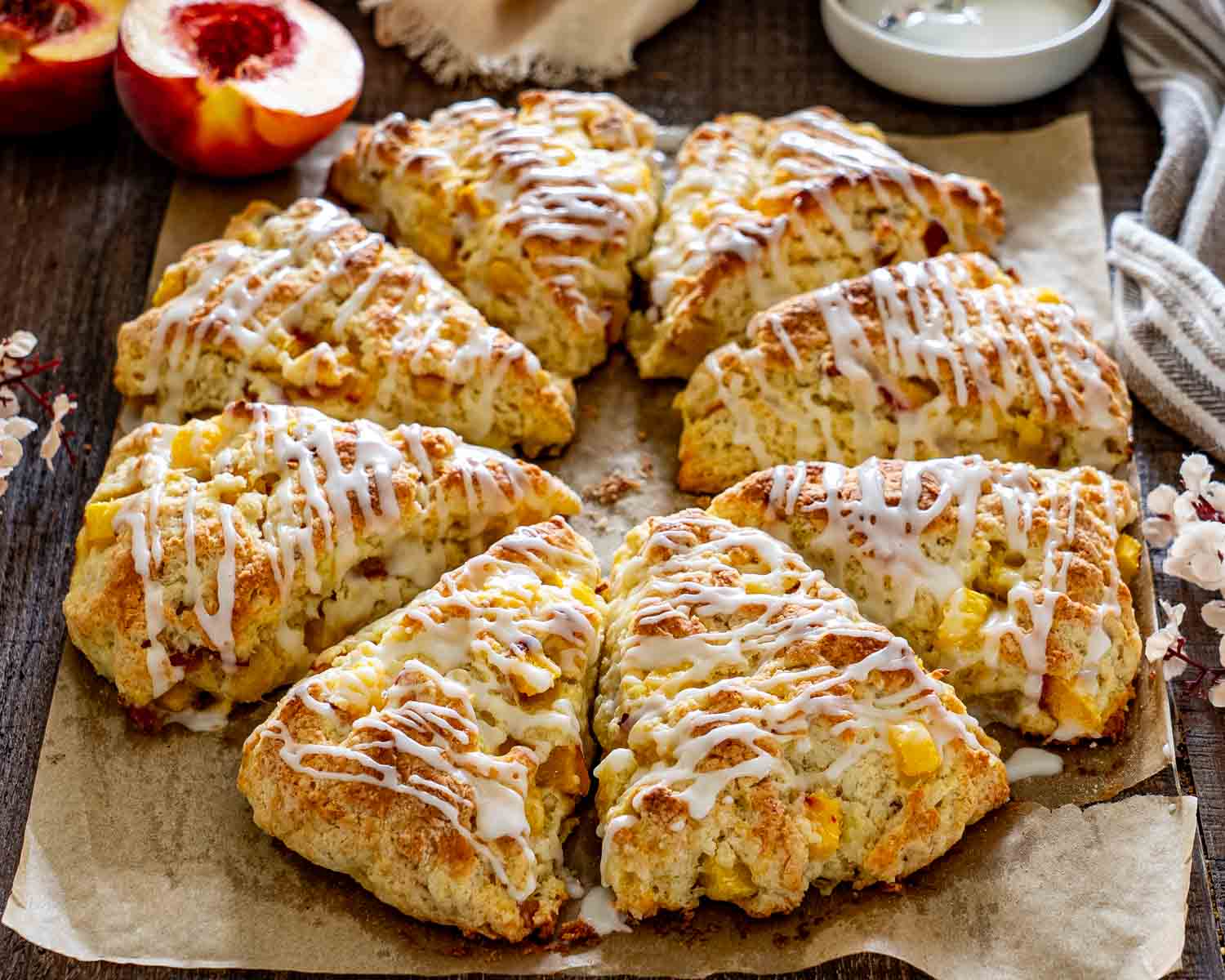 Golden peach scones drizzled with vanilla glaze and arranged on parchment beside fresh peaches and delicate white flowers.