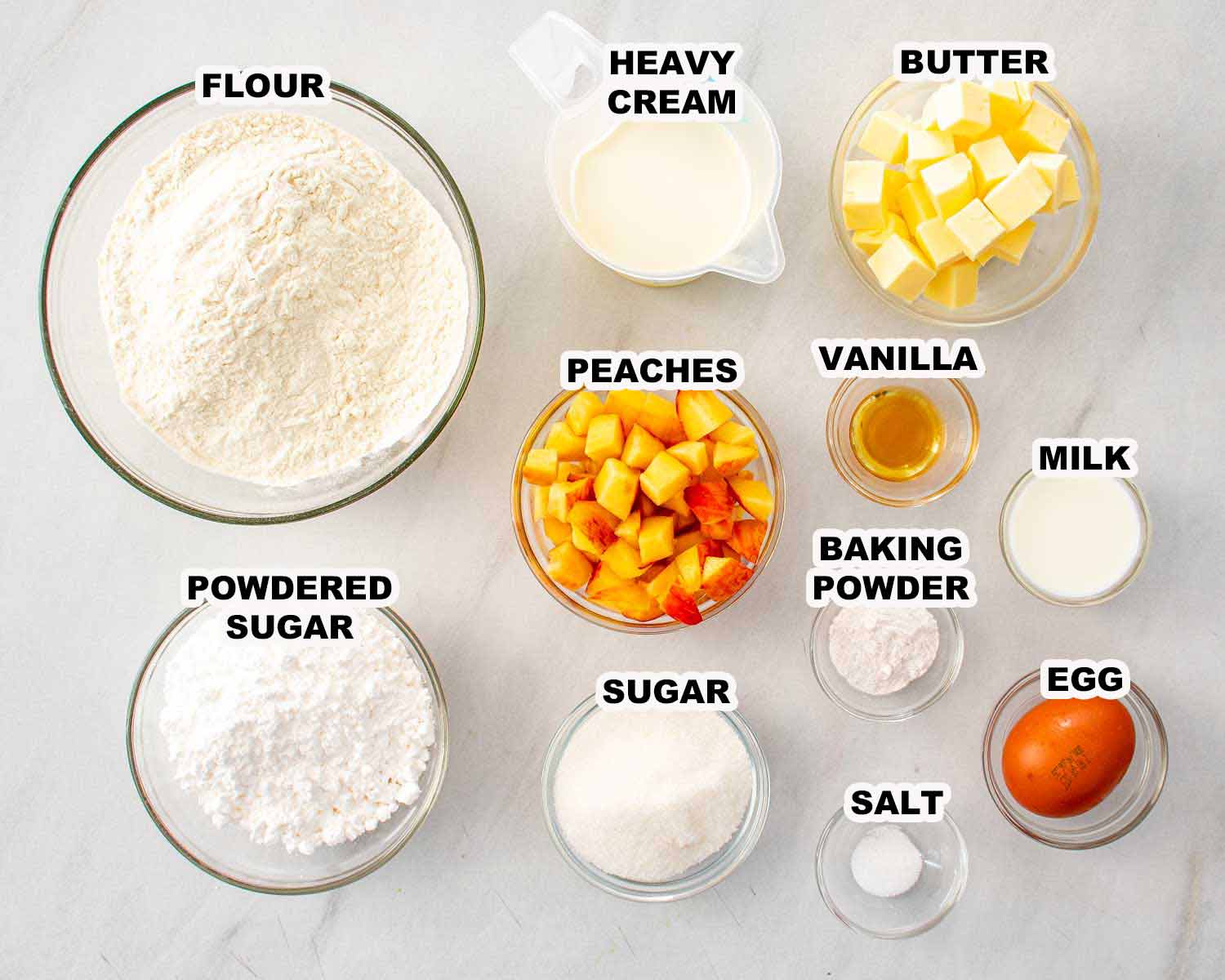 Overhead view of labeled peach scone ingredients including flour, peaches, butter, cream, sugar, milk, egg, and vanilla.