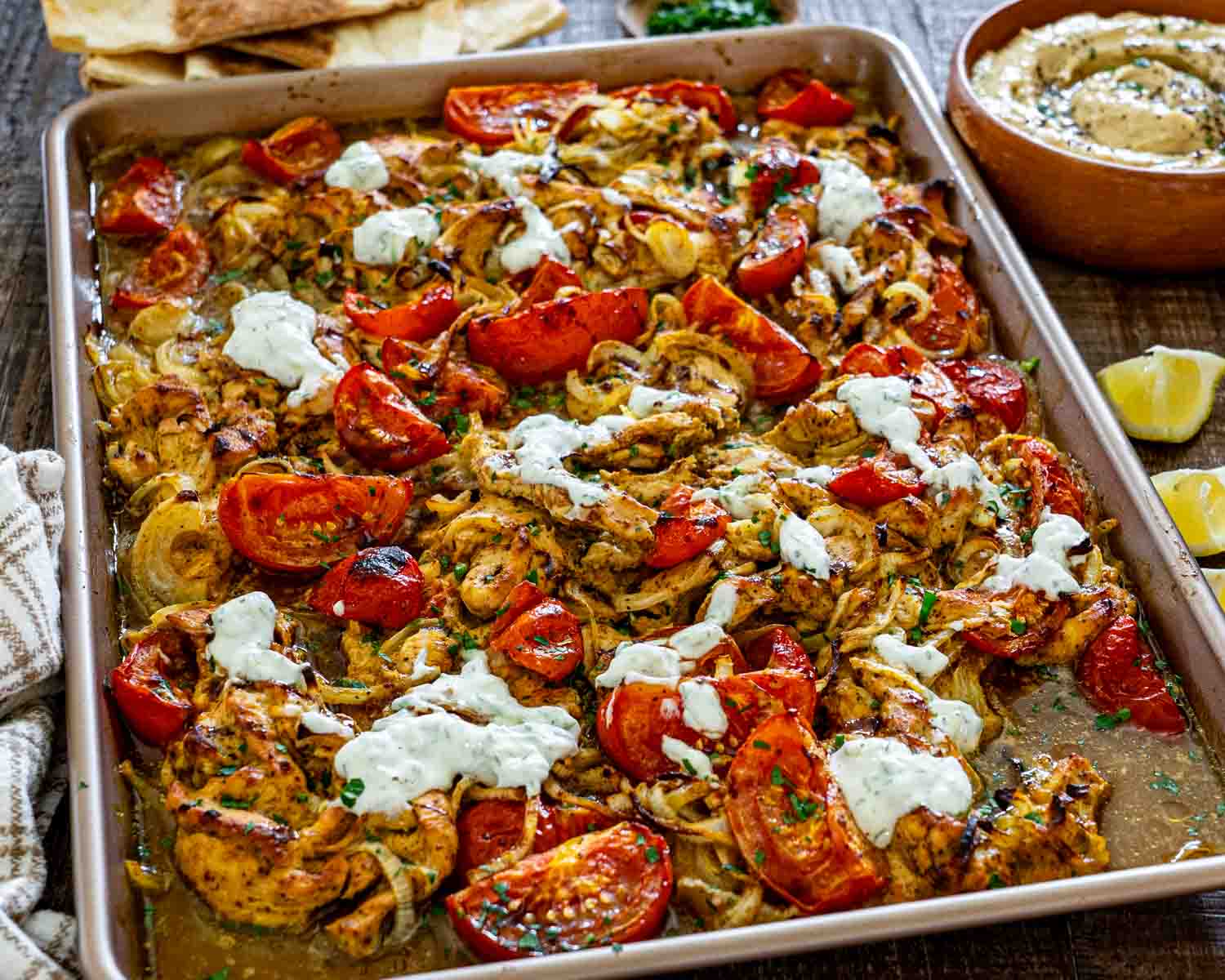 Angled view of sheet pan chicken shawarma with caramelized onions, roasted tomatoes, parsley, yogurt sauce, hummus, and lemons.