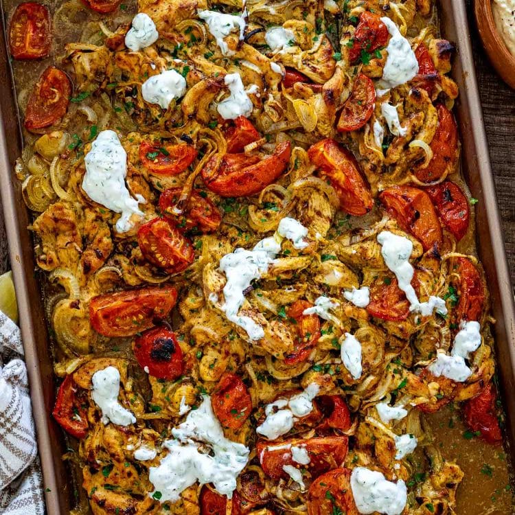Overhead view of sheet pan chicken shawarma with roasted tomatoes, onions, parsley, and dollops of creamy yogurt sauce.