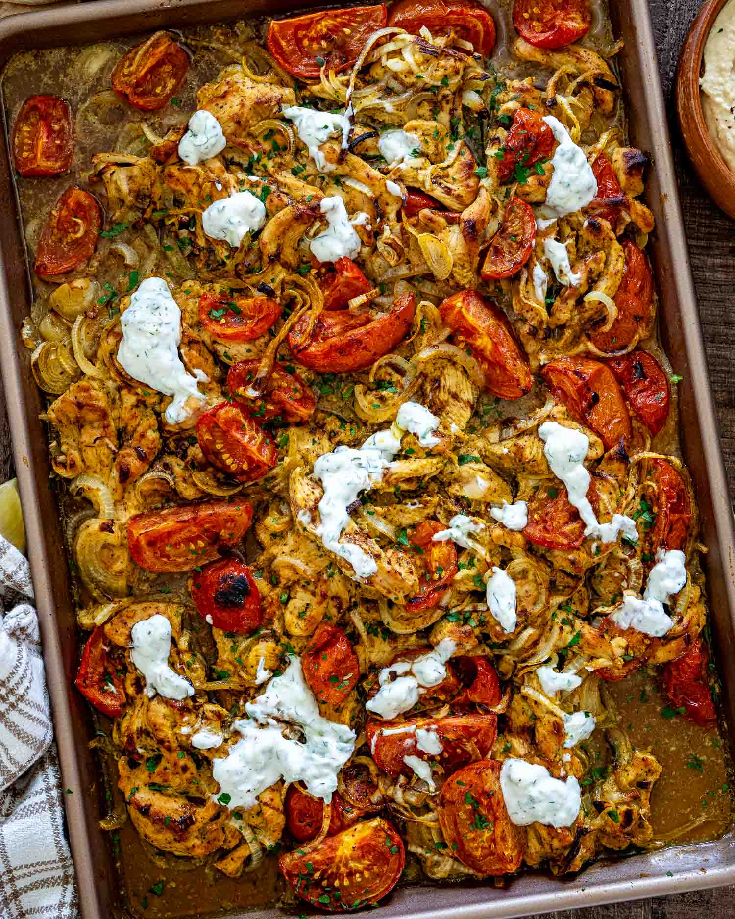 Overhead view of sheet pan chicken shawarma with roasted tomatoes, onions, parsley, and dollops of creamy yogurt sauce.