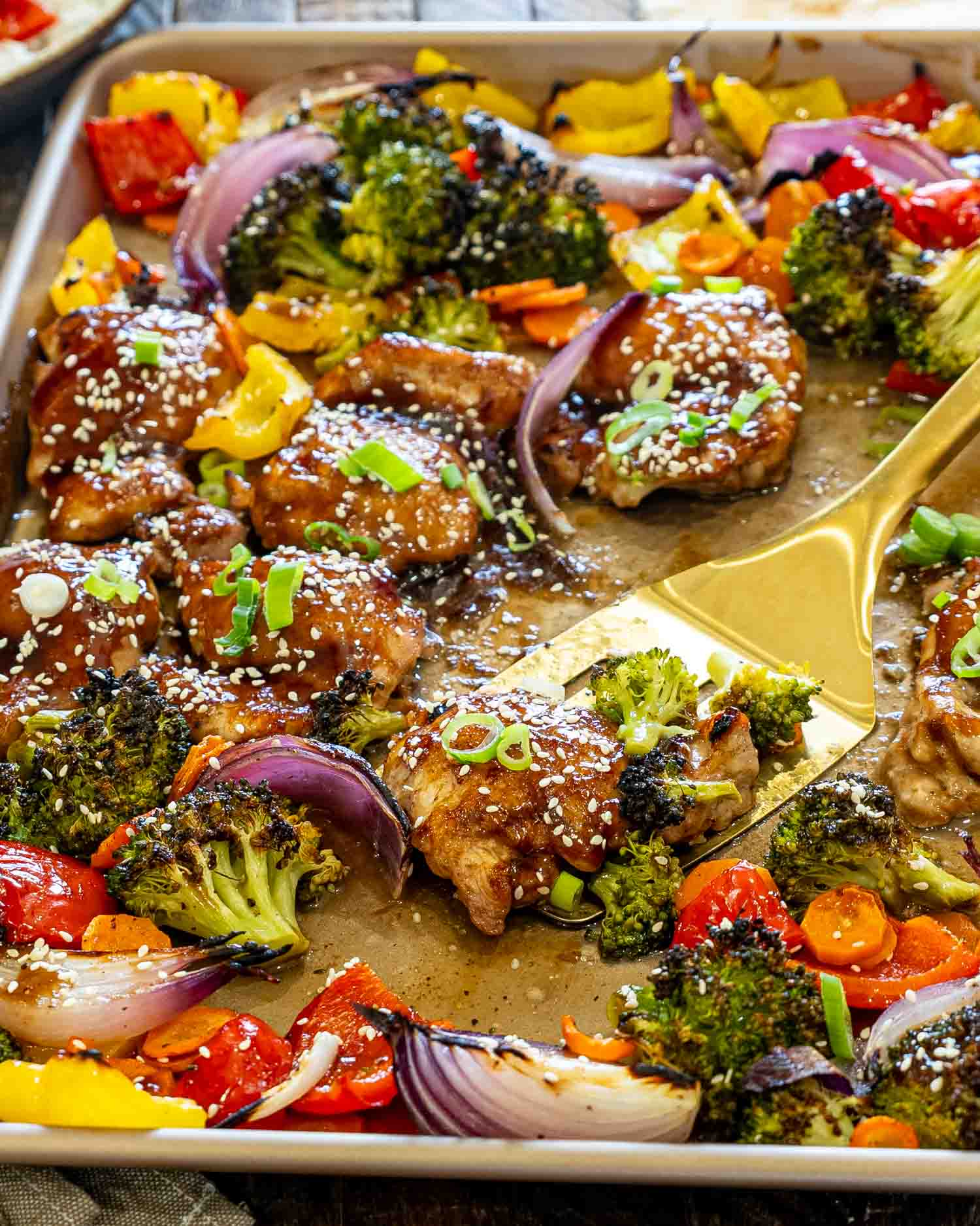Golden spatula lifting a glazed teriyaki chicken thigh with roasted broccoli, peppers, onions, carrots, sesame seeds, and green onions.