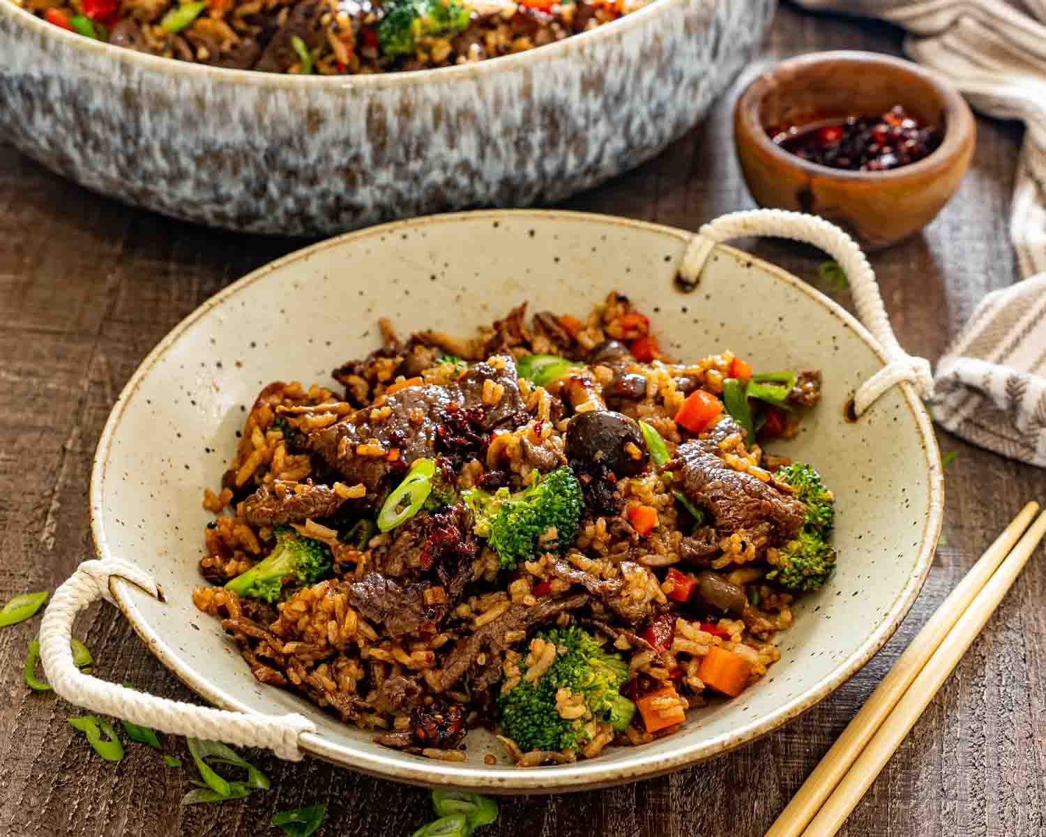 Rustic bowl filled with sticky beef fried rice served alongside chopsticks, featuring tender beef, broccoli, mushrooms, peppers, and carrots.