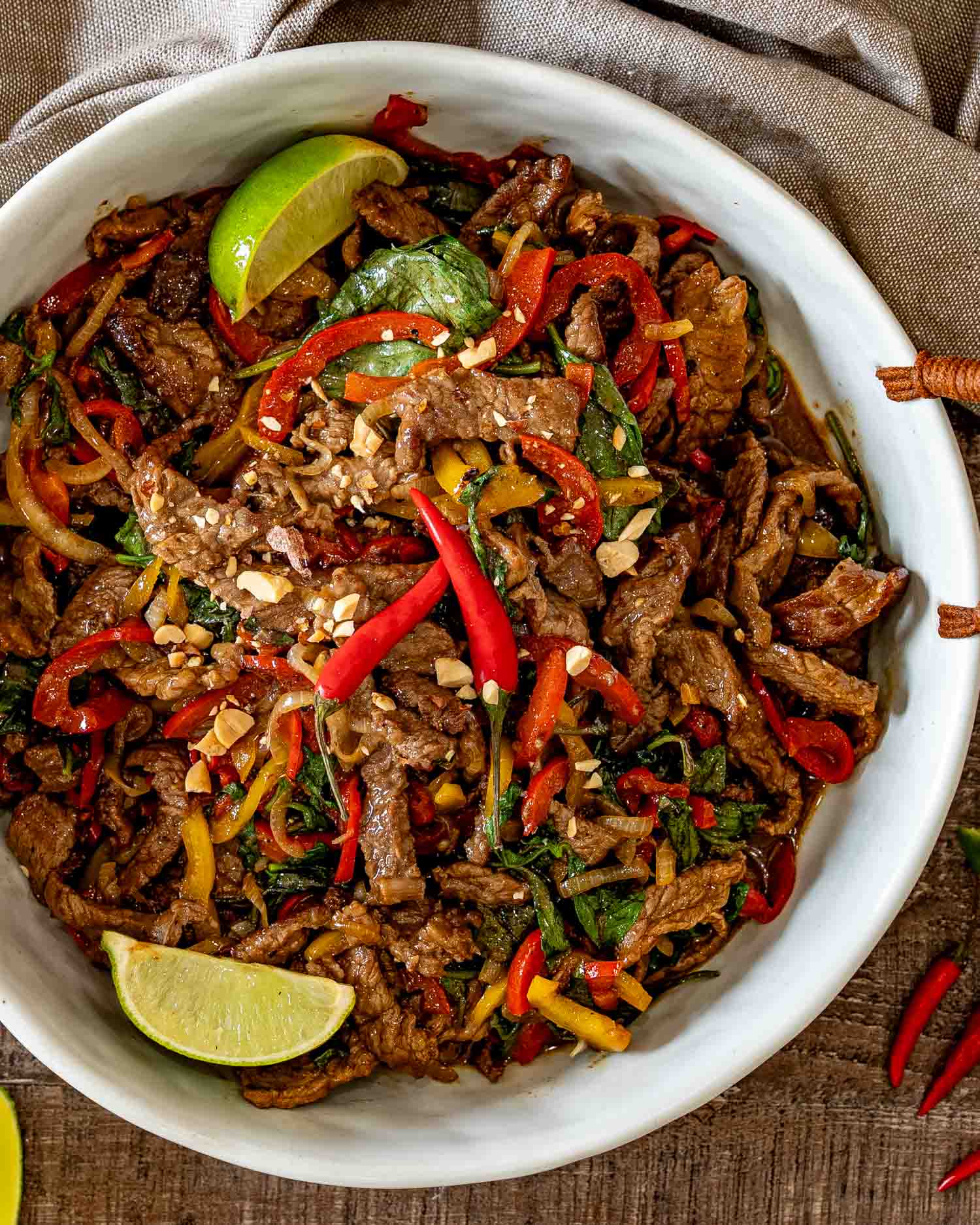 Overhead serving bowl filled with Thai basil beef, colorful peppers, fresh basil, chopped peanuts, chilies, and lime wedges.