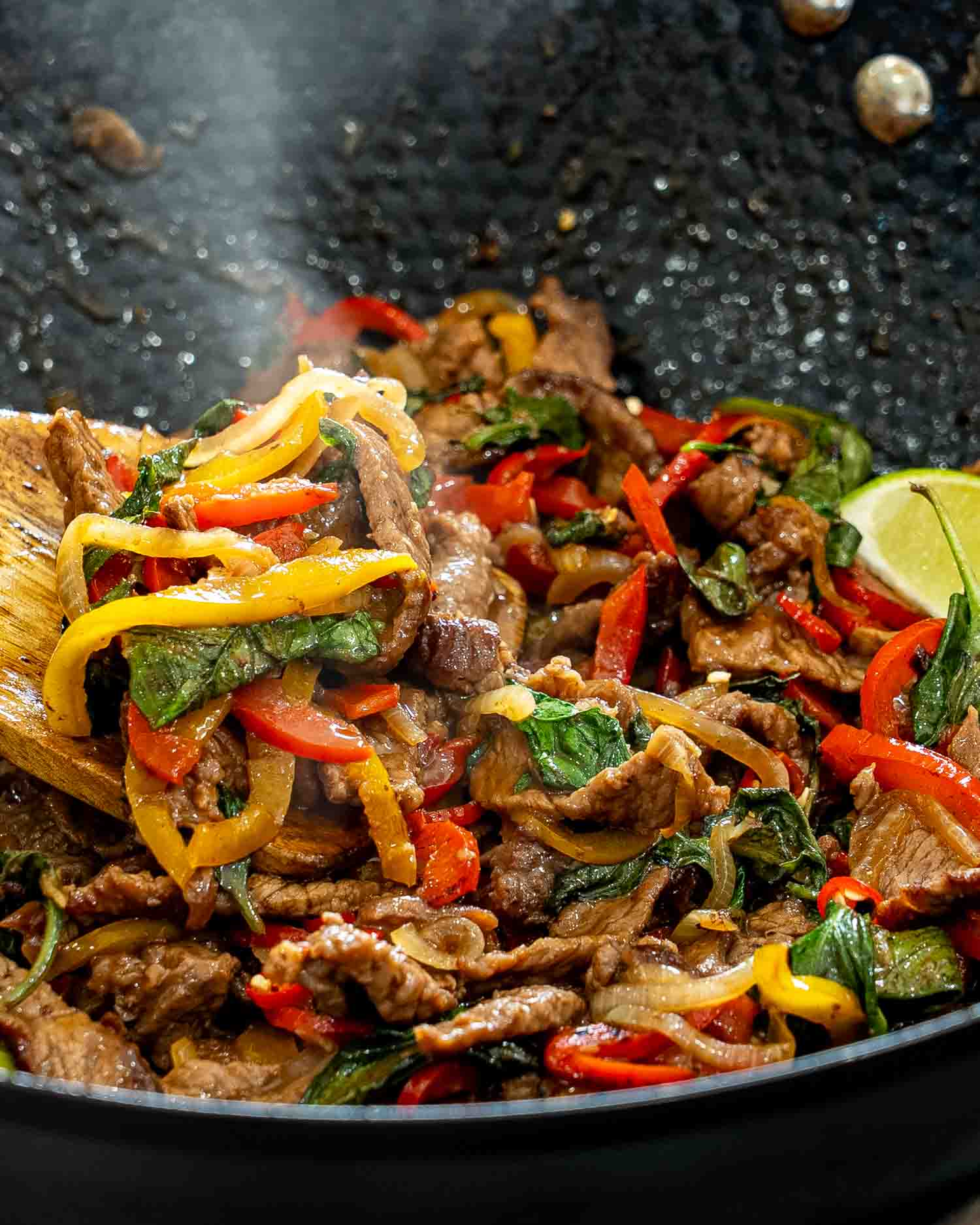 A wooden spoon lifts steaming Thai basil beef with red and yellow peppers from the hot skillet.