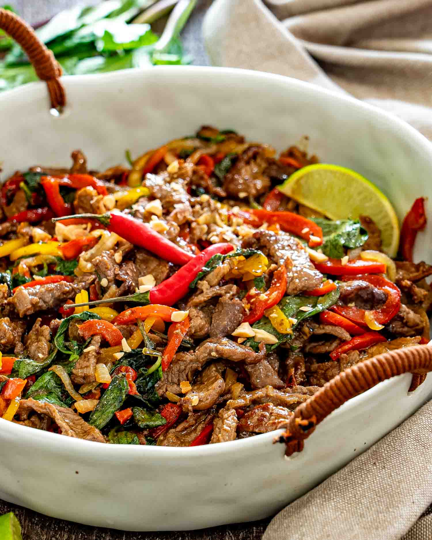 Close-up bowl of Thai basil beef with colorful peppers, red chilies, rice, chopped peanuts, and fresh lime wedges.