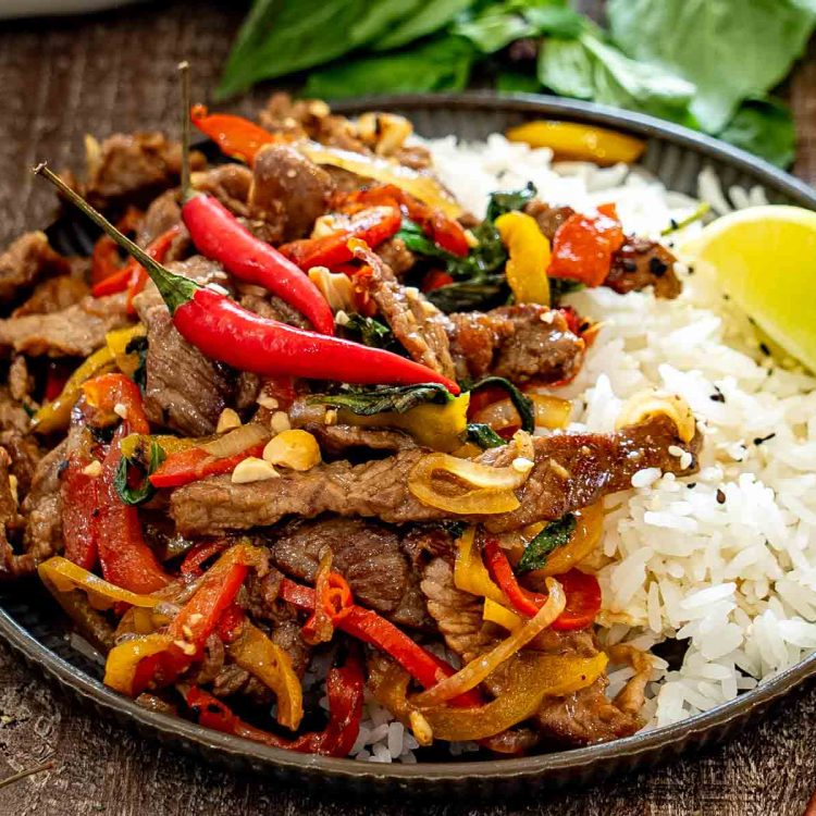 Thai basil beef with peppers and chilies is served beside fluffy white rice, chopped peanuts, and fresh lime.