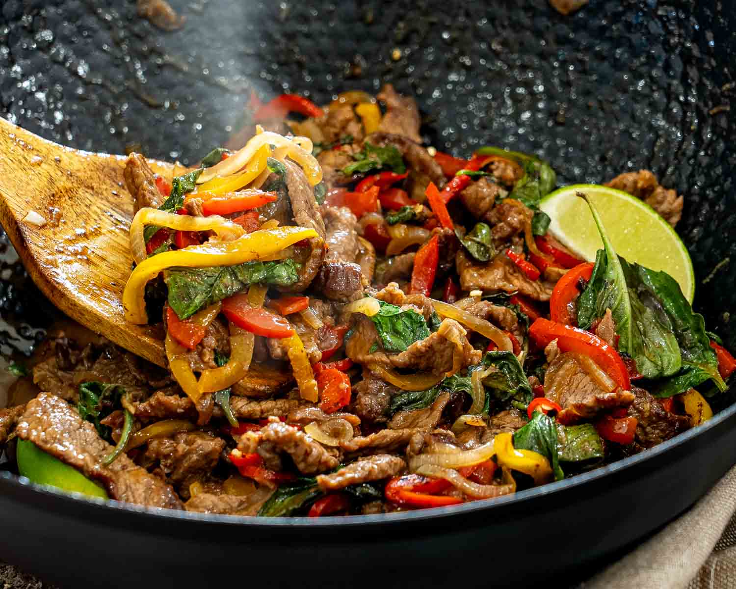 Steaming Thai basil beef with red and yellow peppers, onions, and fresh basil is lifted from a skillet.