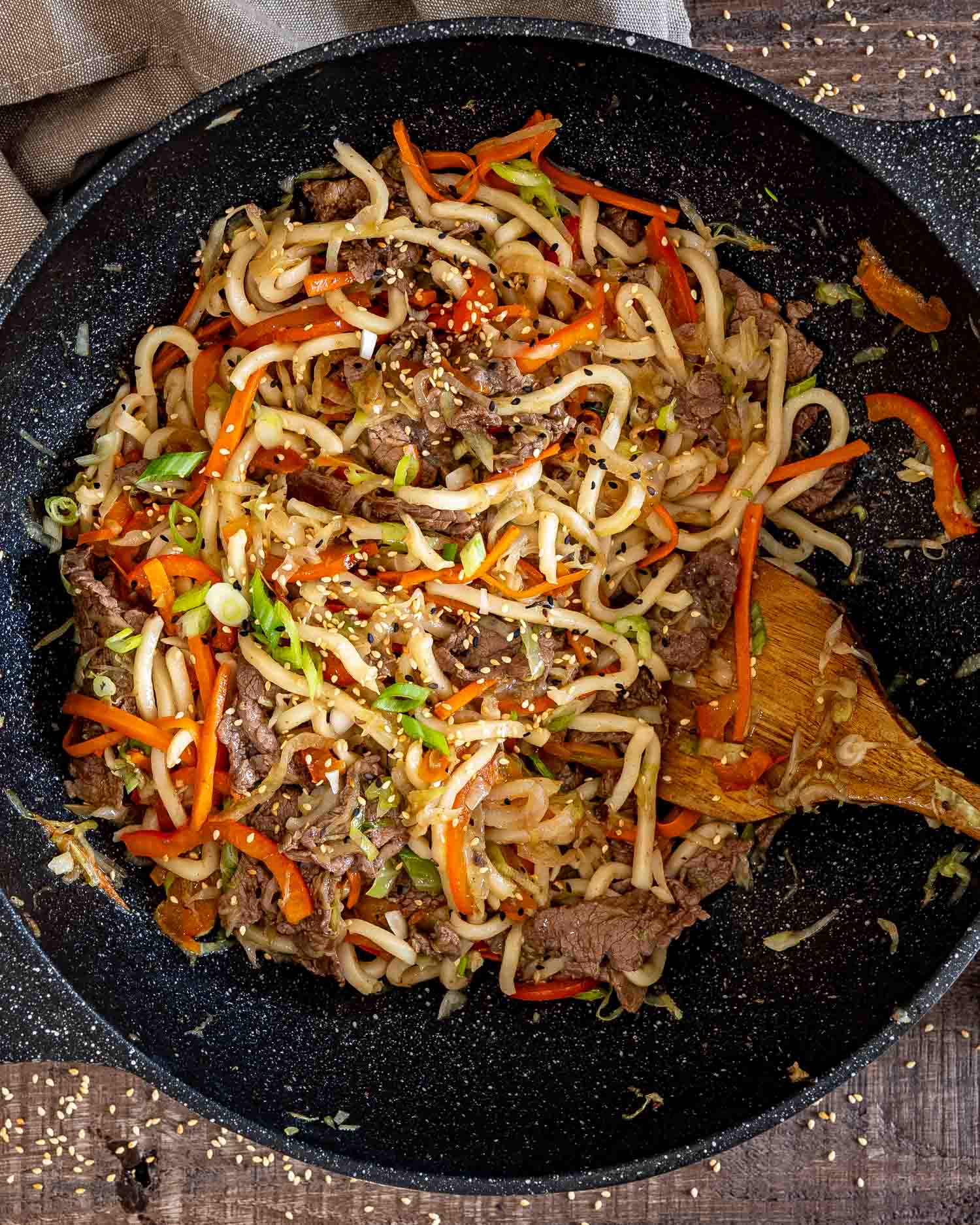Overhead view of beef yaki udon with carrots, peppers, cabbage, and sesame seeds in a black speckled wok.