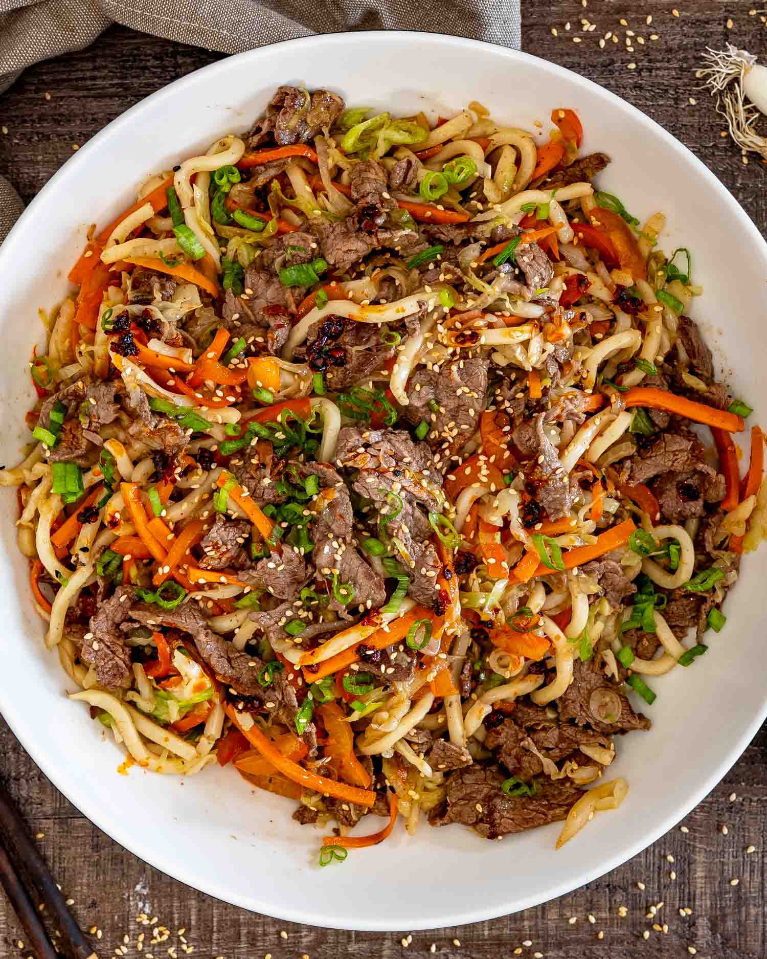 Overhead view of a white serving bowl filled with beef yaki udon, colorful vegetables, sesame seeds, and green onions.