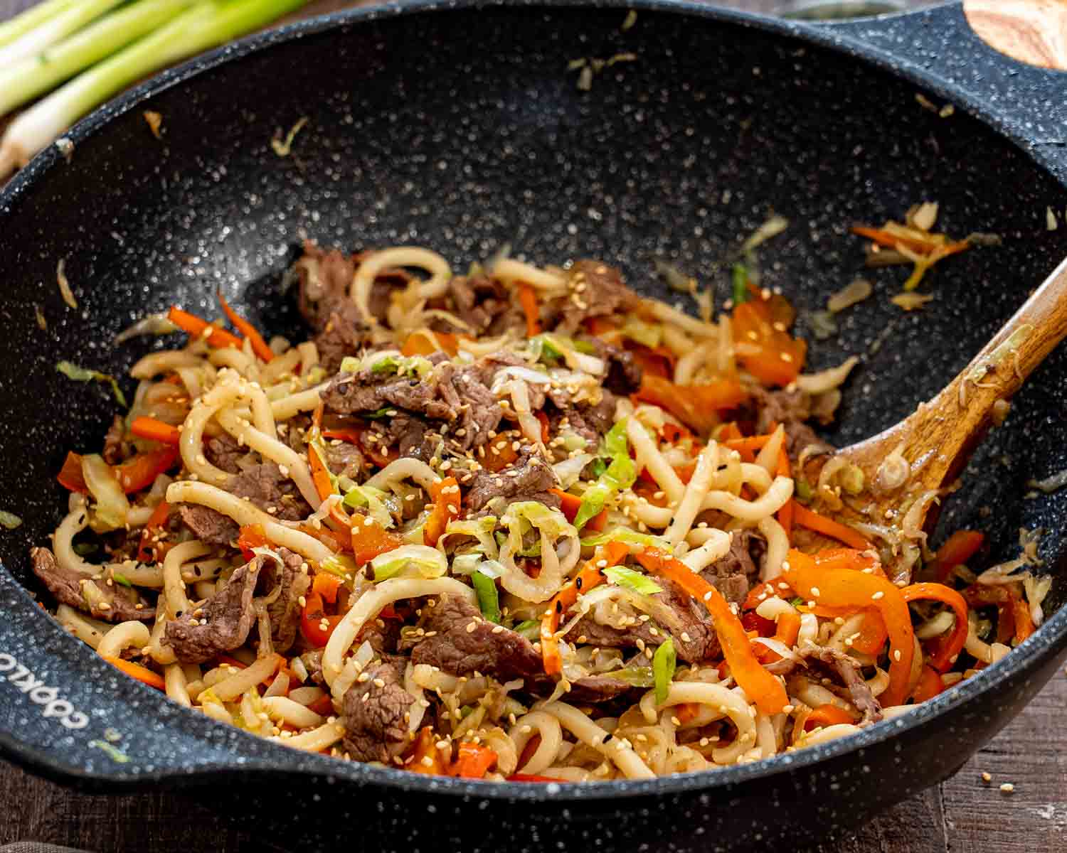 Angled close view of beef yaki udon with sesame seeds and vegetables in a wok beside a wooden spatula.