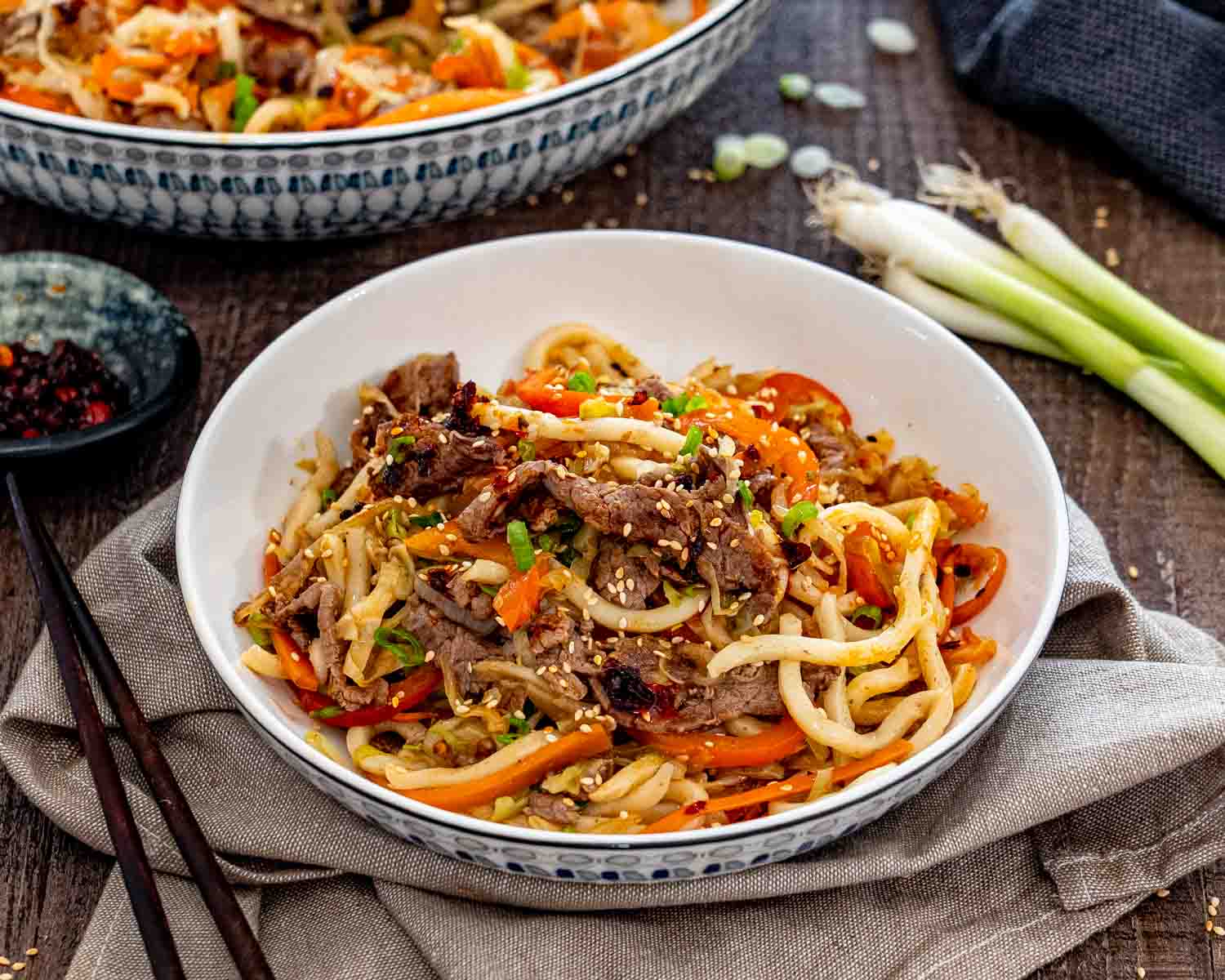 Beef yaki udon served in a patterned bowl on beige linen, with chopsticks and green onions arranged nearby.