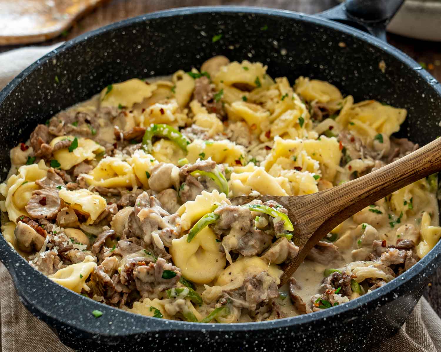 Wooden spoon lifts creamy cheesesteak tortellini with sliced steak, mushrooms, peppers, Parmesan, parsley, and crushed red pepper.