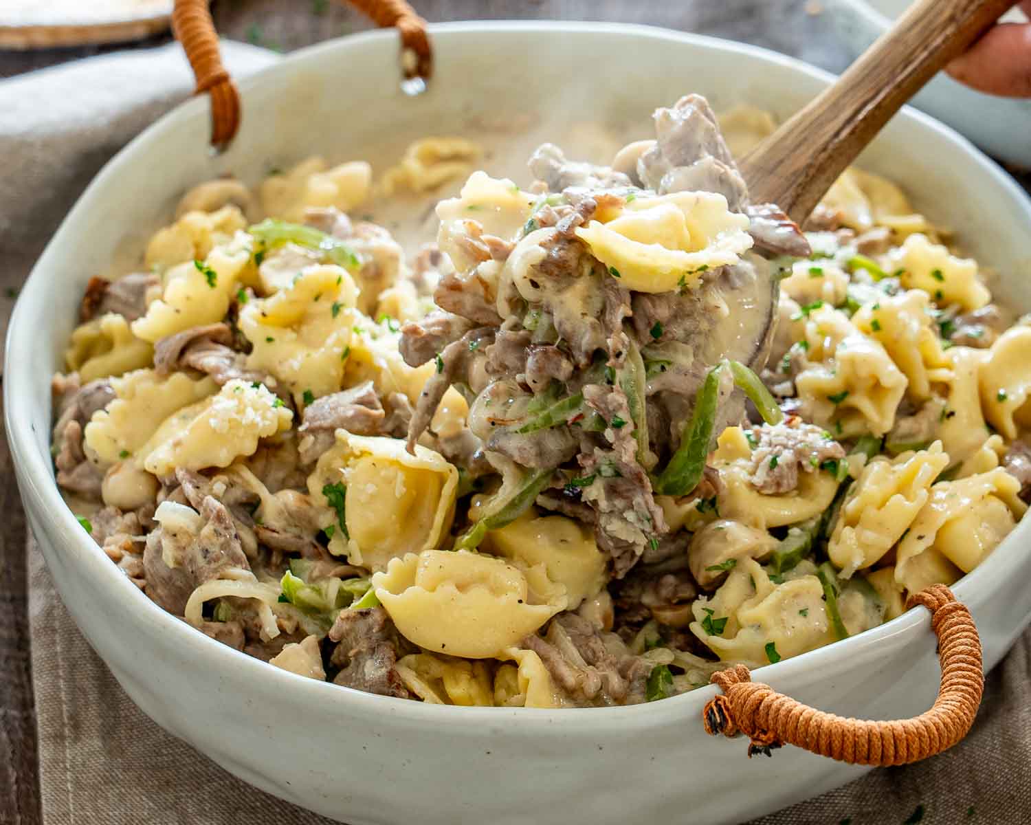 Wooden spoon lifts steak, tortellini, mushrooms, and peppers coated in creamy cheese sauce from a white casserole dish.