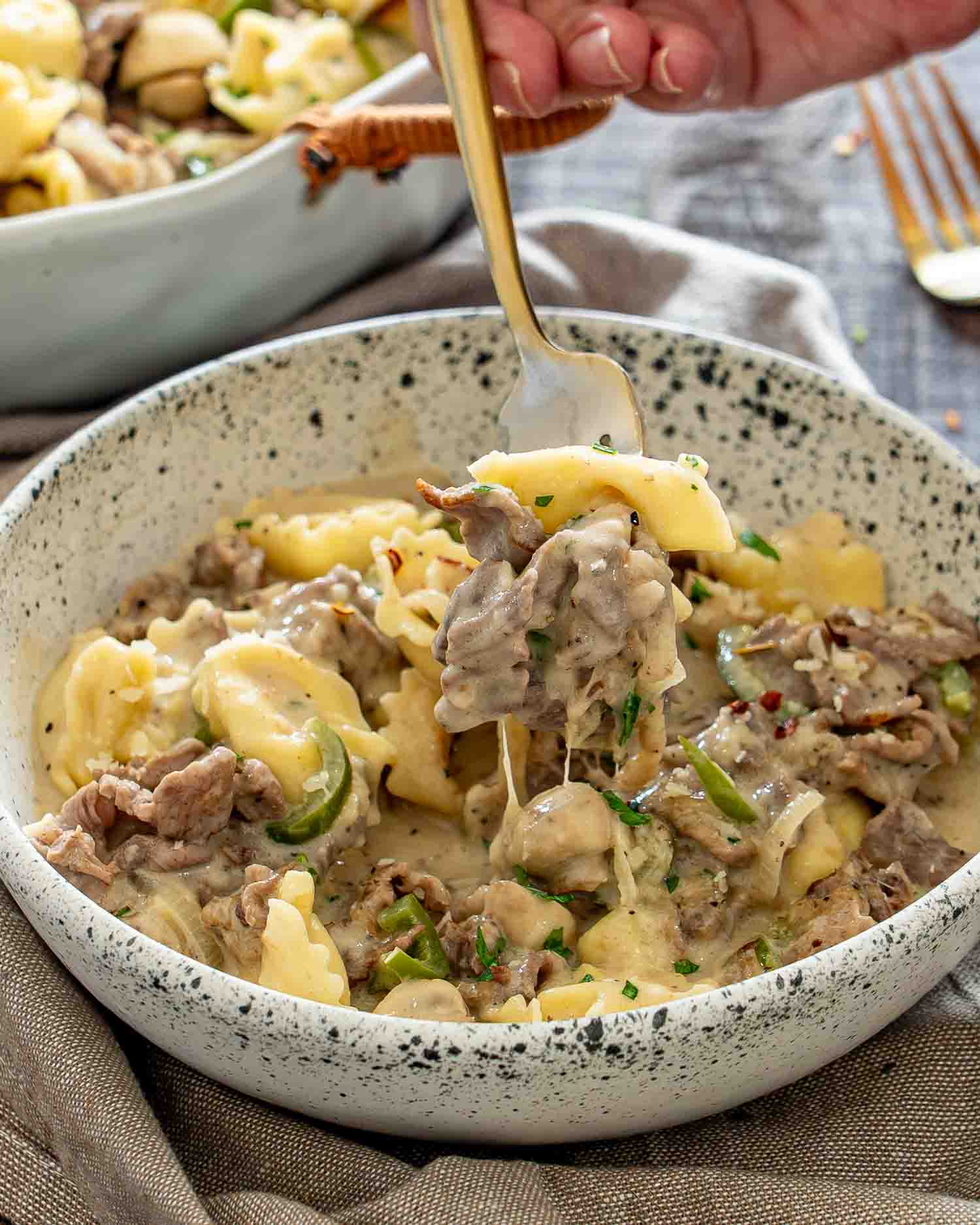 A fork lifts a creamy bite of cheesesteak tortellini with tender steak from a speckled serving bowl.
