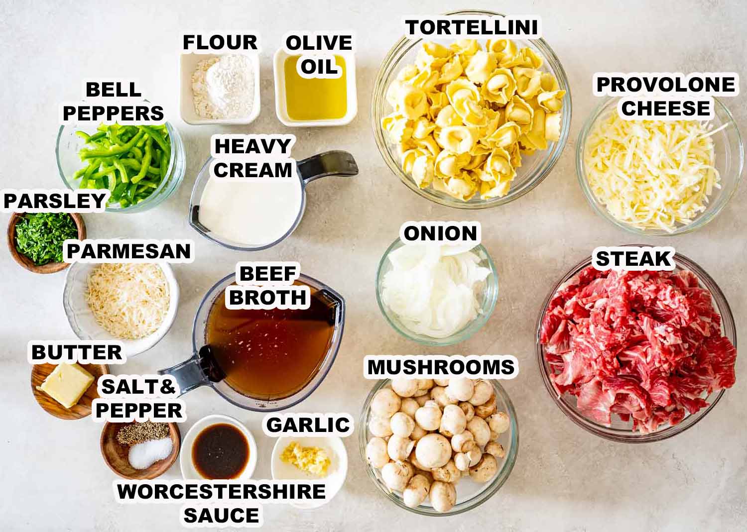 Overhead arrangement of labeled ingredients needed to make creamy cheesesteak tortellini, including steak, cheeses, vegetables, and seasonings.