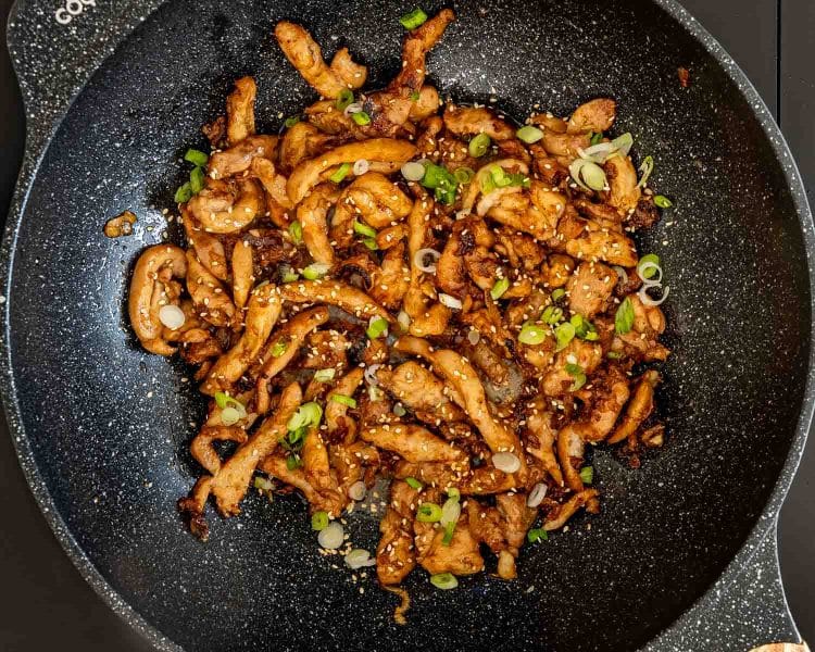 Overhead skillet filled with browned chicken bulgogi, scattered sesame seeds, and sliced green onions ready for serving.