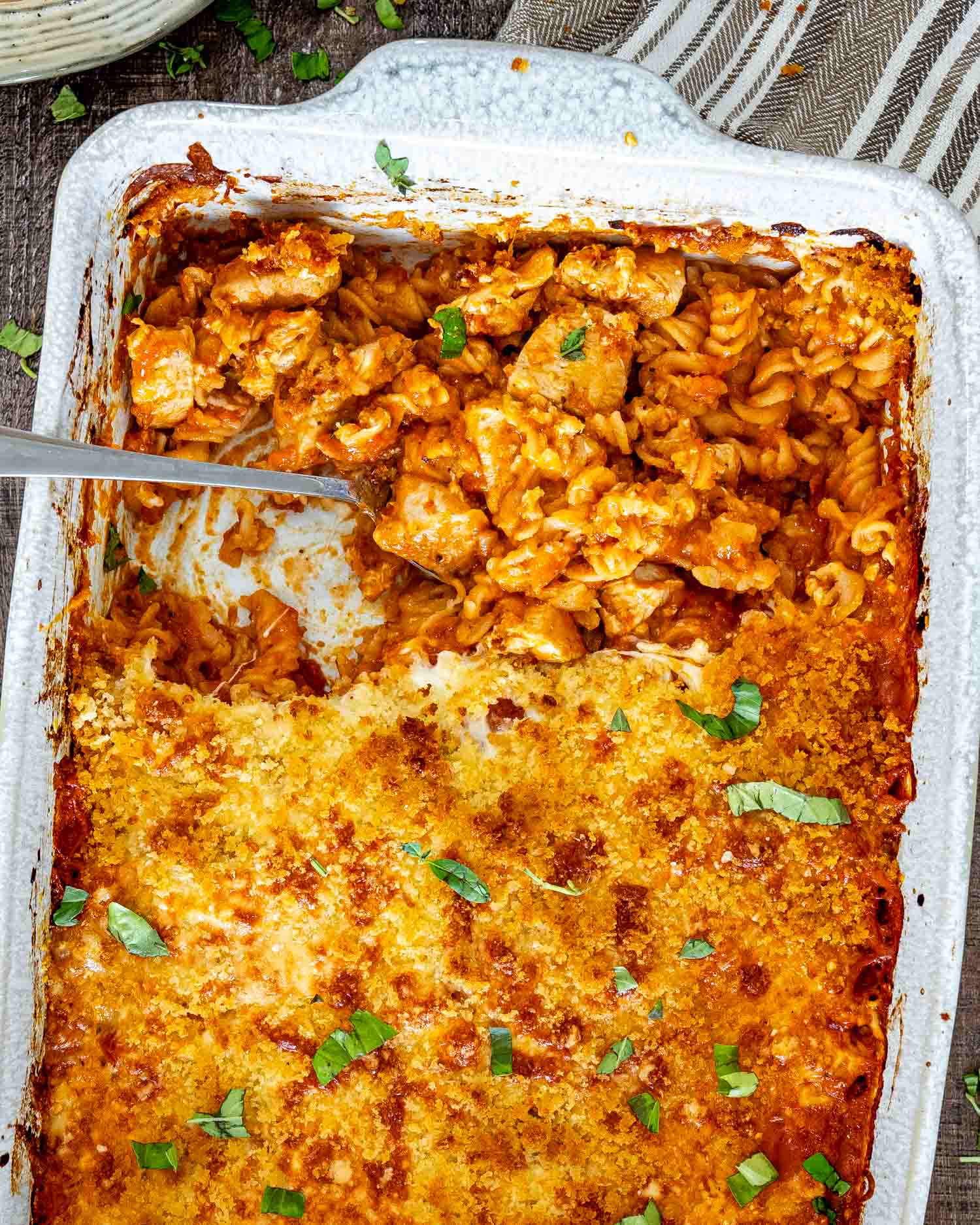 A spoon lifts cheesy chicken and saucy rotini from the golden breadcrumb-topped casserole, garnished with chopped fresh basil.