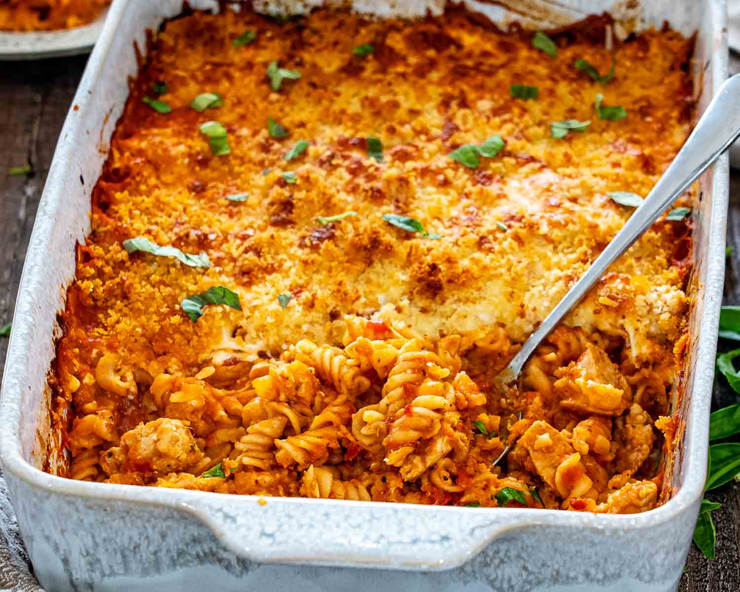 Close view of baked chicken Parmesan casserole with golden breadcrumbs, melted cheese, rotini, chicken pieces, and basil.