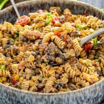 Spoonful of creamy cowboy pasta salad lifted from a large bowl, showing beef, bacon, beans, corn, and tomatoes.