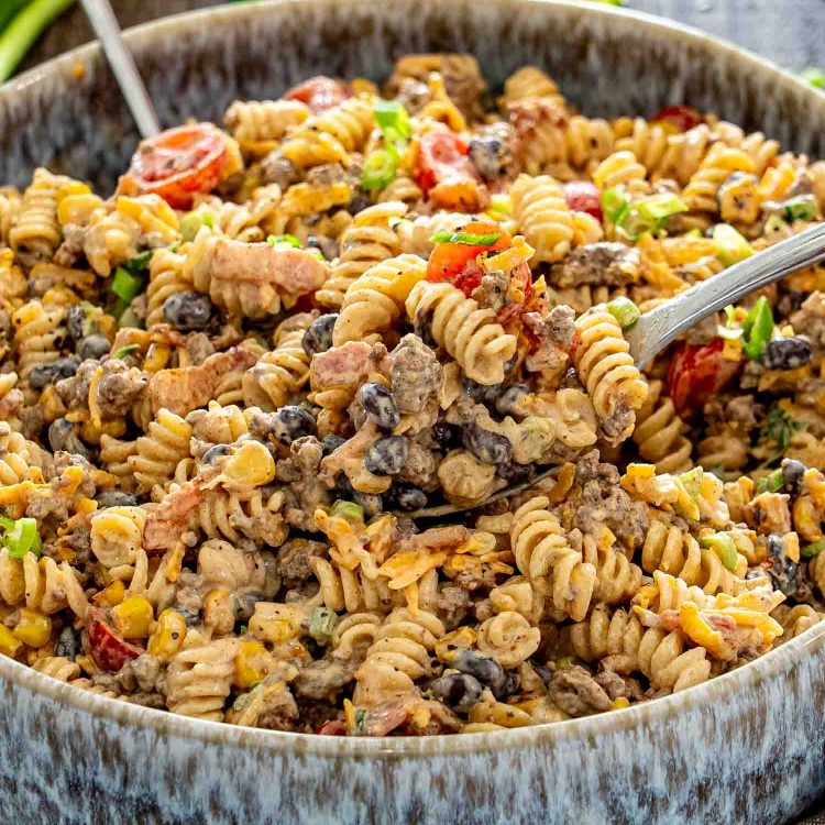 Spoonful of creamy cowboy pasta salad lifted from a large bowl, showing beef, bacon, beans, corn, and tomatoes.