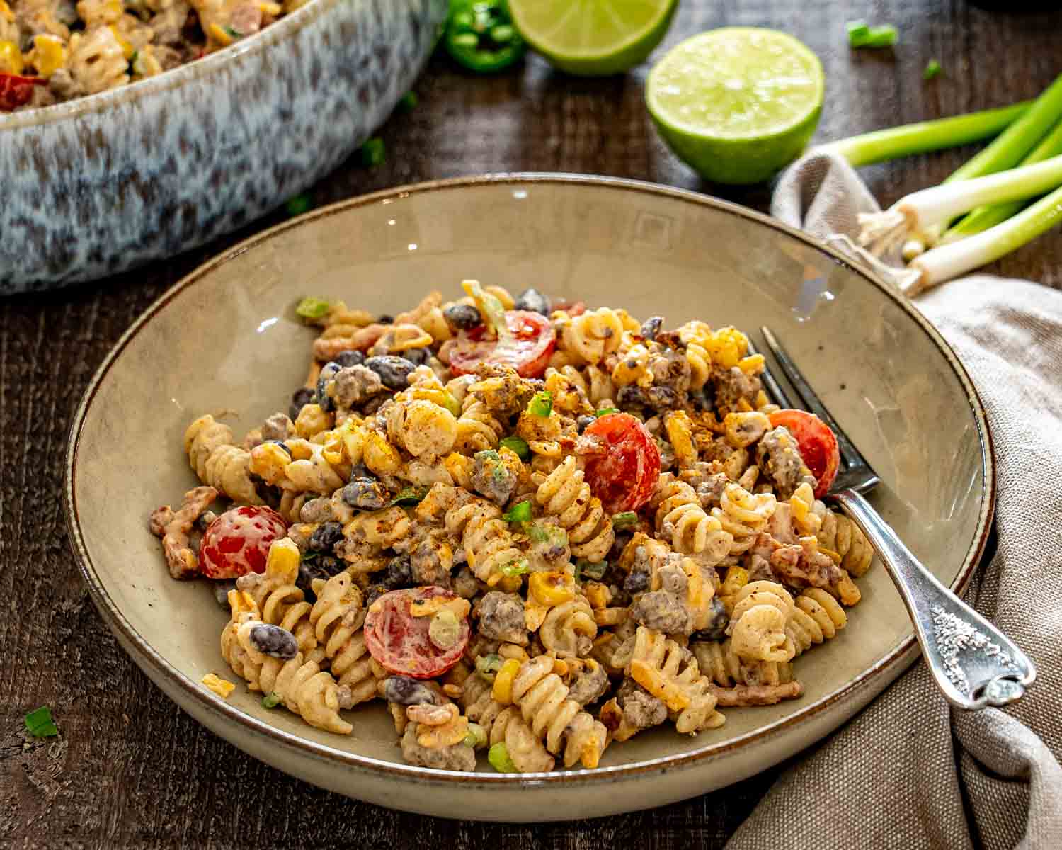 Cowboy pasta salad served in a shallow bowl with lime, green onions, and the larger serving dish behind it.