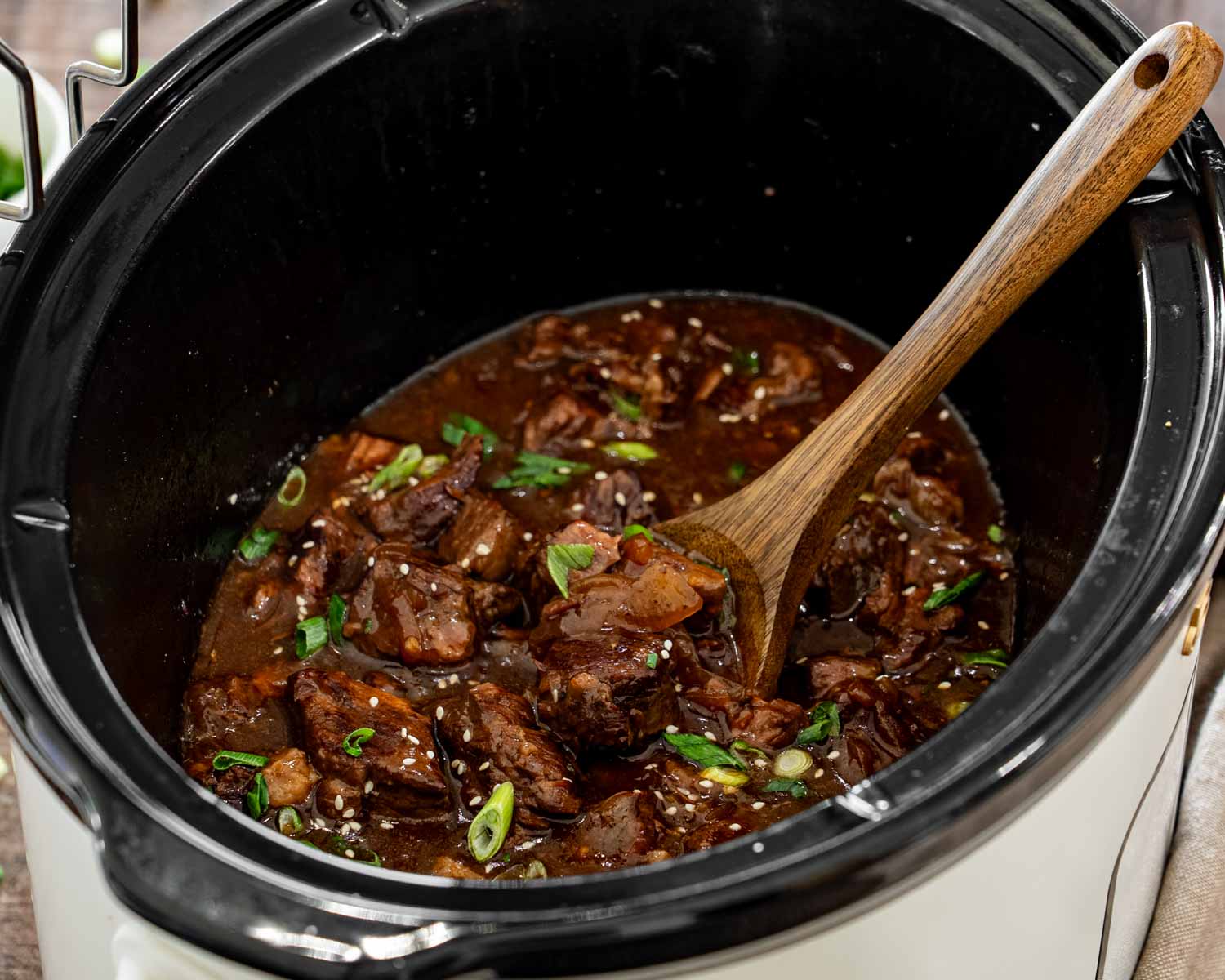 Tender Asian braised beef in a slow cooker with glossy sauce, sesame seeds, green onions, and a wooden spoon.