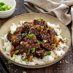 Asian braised beef piled over steamed white rice, garnished with sesame seeds and green onions in a shallow bowl. (