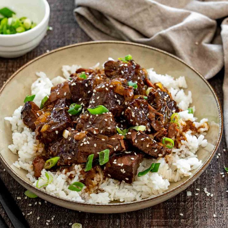 Asian braised beef piled over steamed white rice, garnished with sesame seeds and green onions in a shallow bowl. (